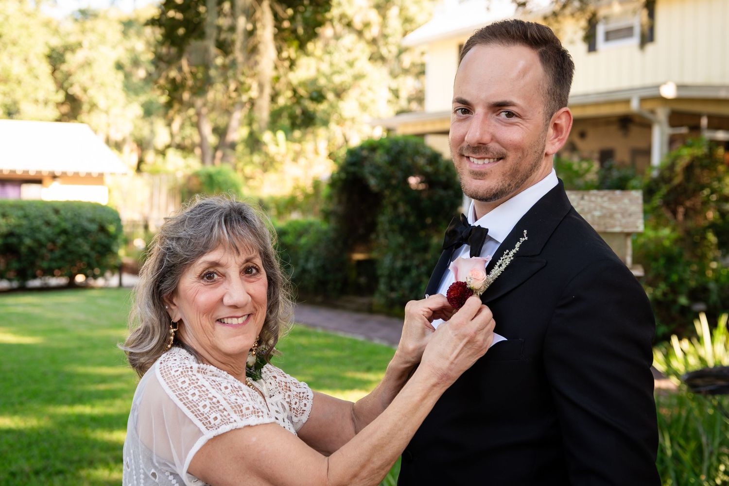 Mother of the groom pinning a boutonniere on her sons tuxedo lapel outdoors at a wedding venue.