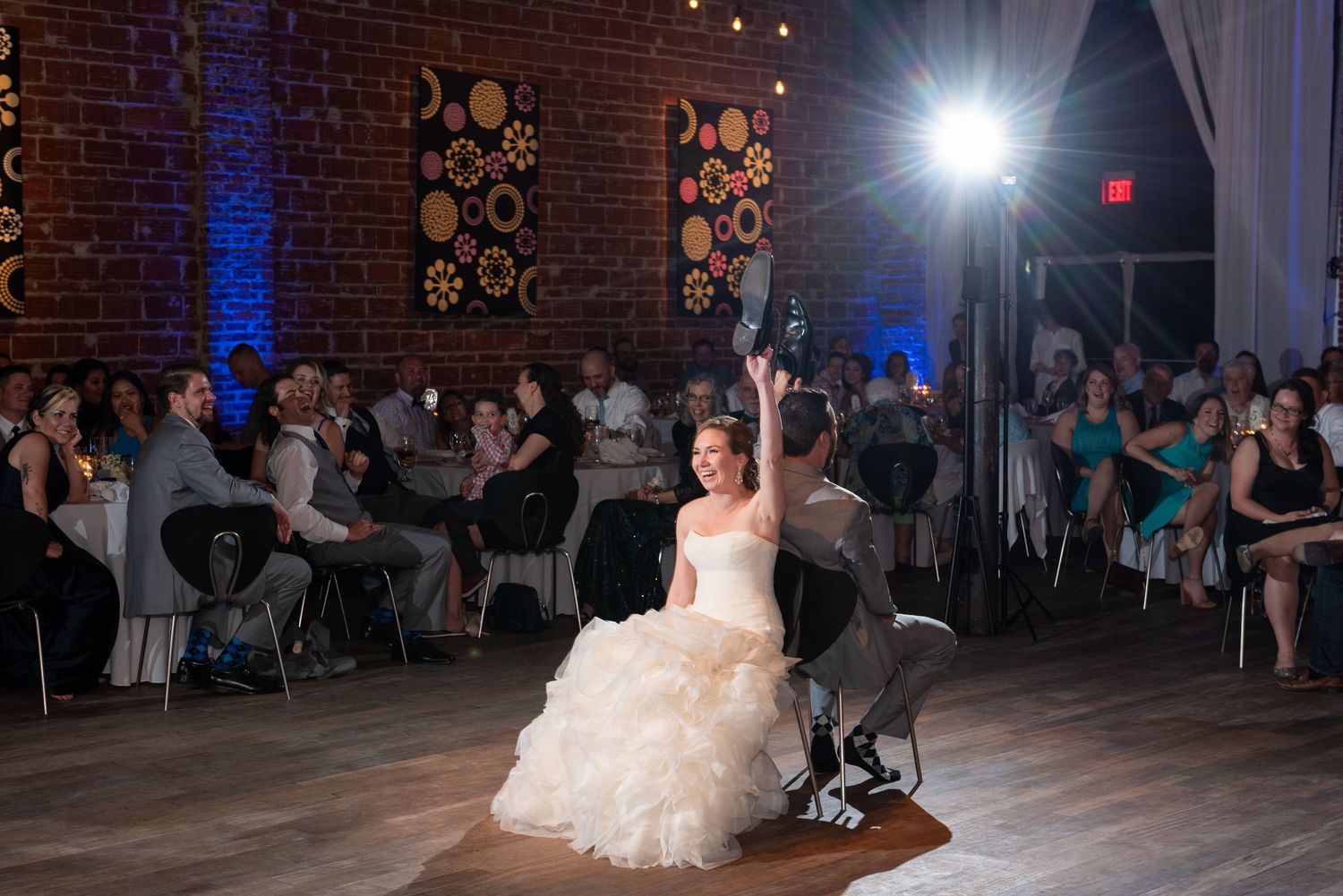 Bride in white ballgown raises hand while seated at wedding reception in brick venue with guests watching.