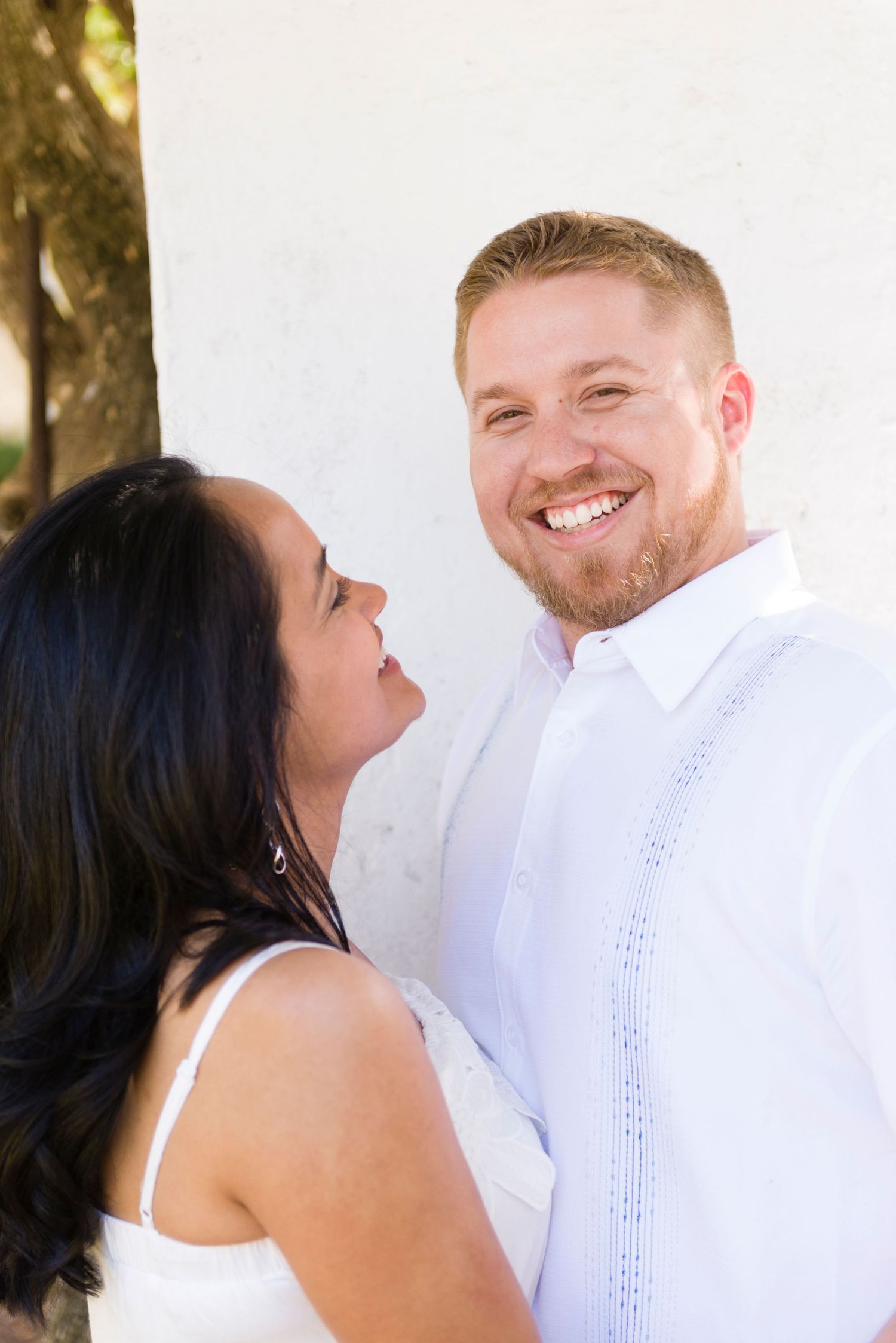 A smiling couple in white attire share a joyful moment against a white wall outdoors.