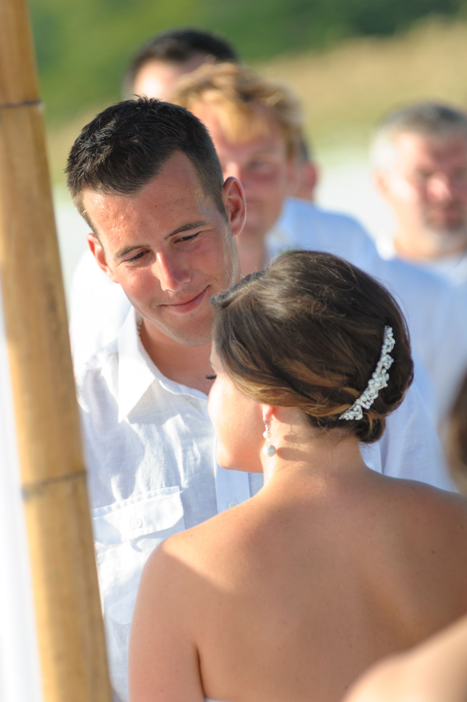 Groom in white shirt smiles lovingly at bride during outdoor beach wedding ceremony.