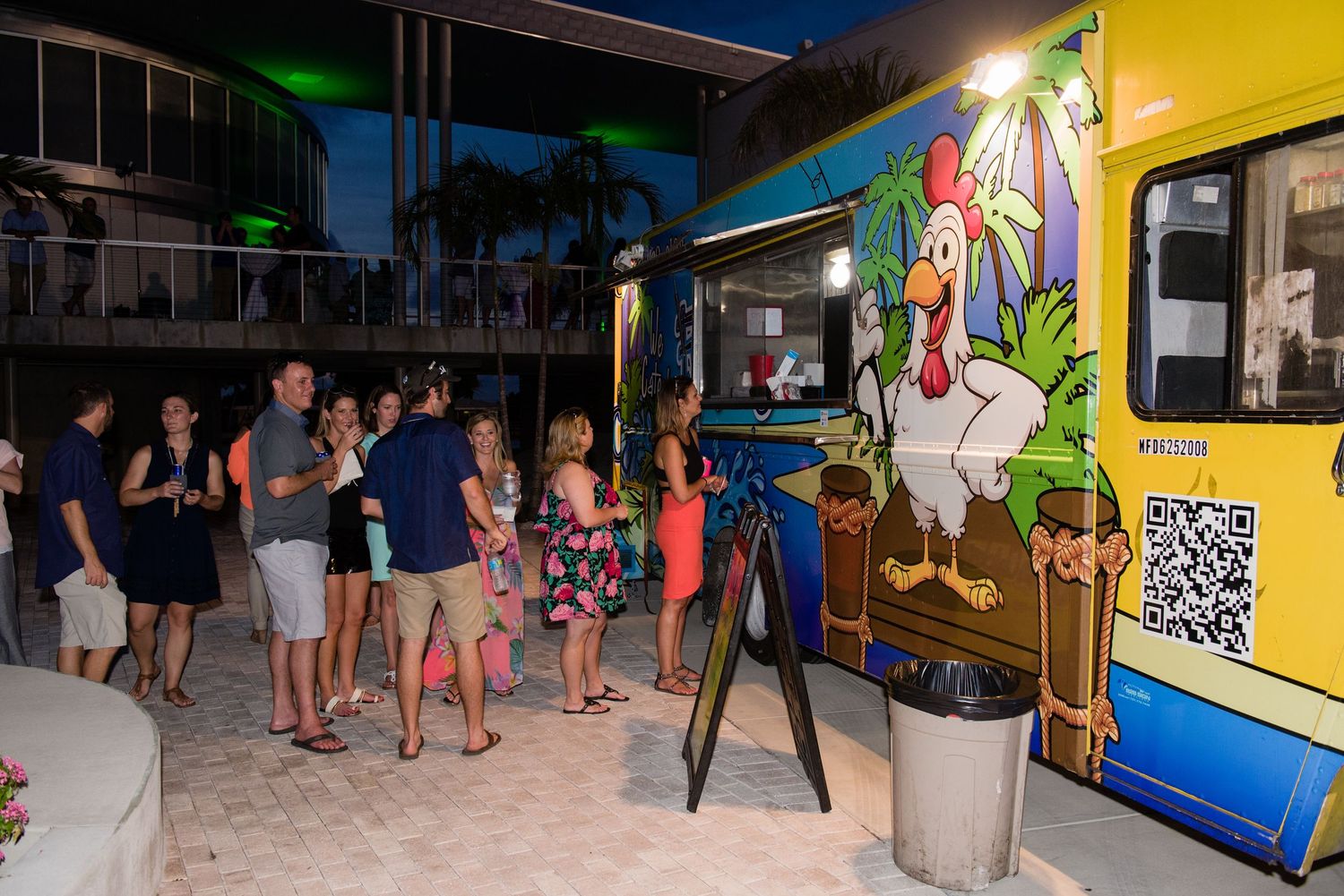 People gather at a colorful food truck with chicken artwork at a tropical outdoor evening event.