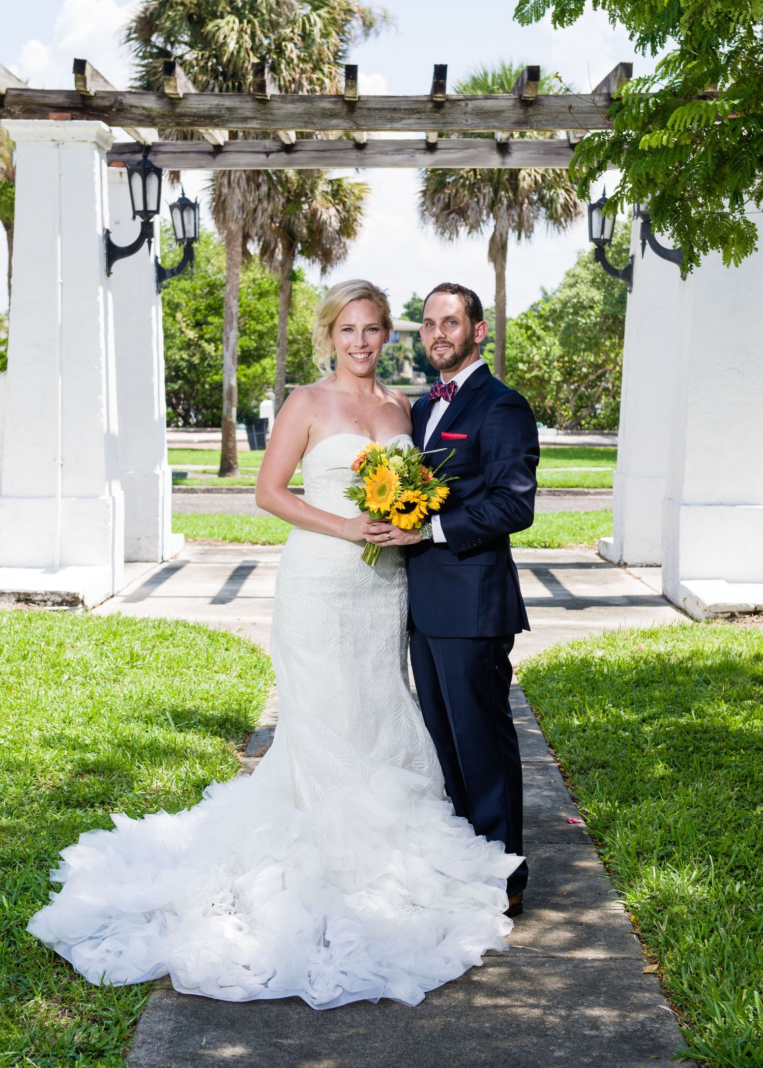 Bride and groom pose together under a white pergola, she holds sunflowers in a strapless gown, he wears a navy tuxedo.
