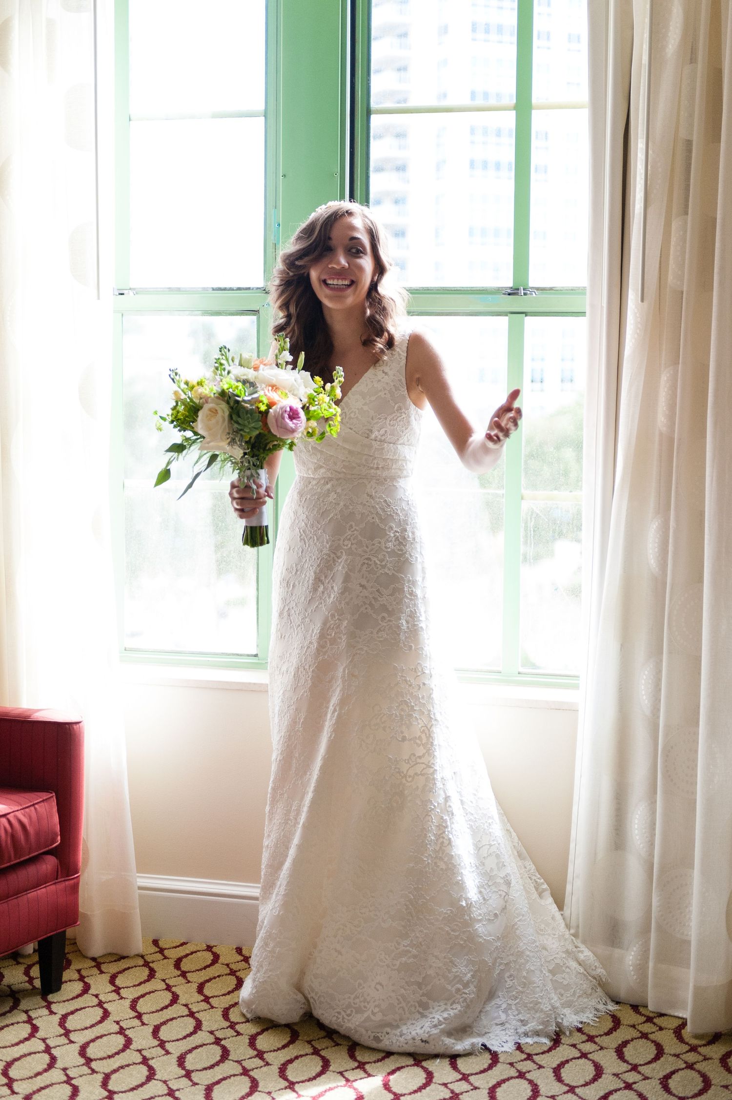 Radiant bride in lace wedding gown holding floral bouquet standing by green-framed window.