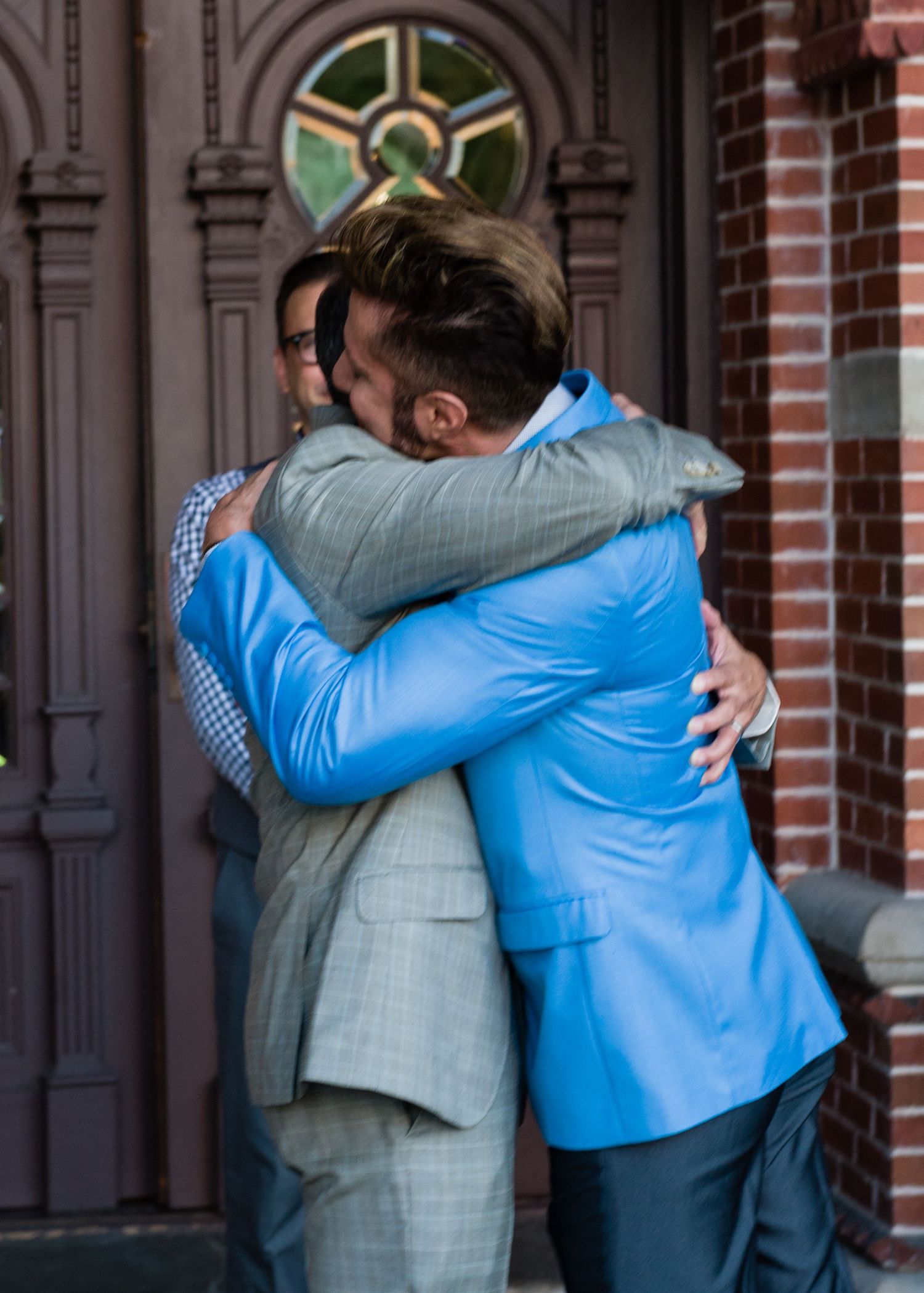 Two men in suits share a warm hug outside a historic brick building with ornate wooden doors.