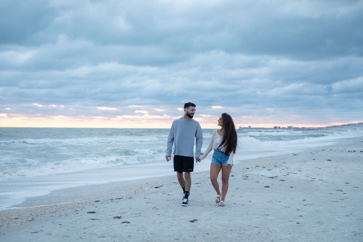 Couple holding hands walking on a sandy beach at sunset with dramatic cloudy skies and ocean waves.