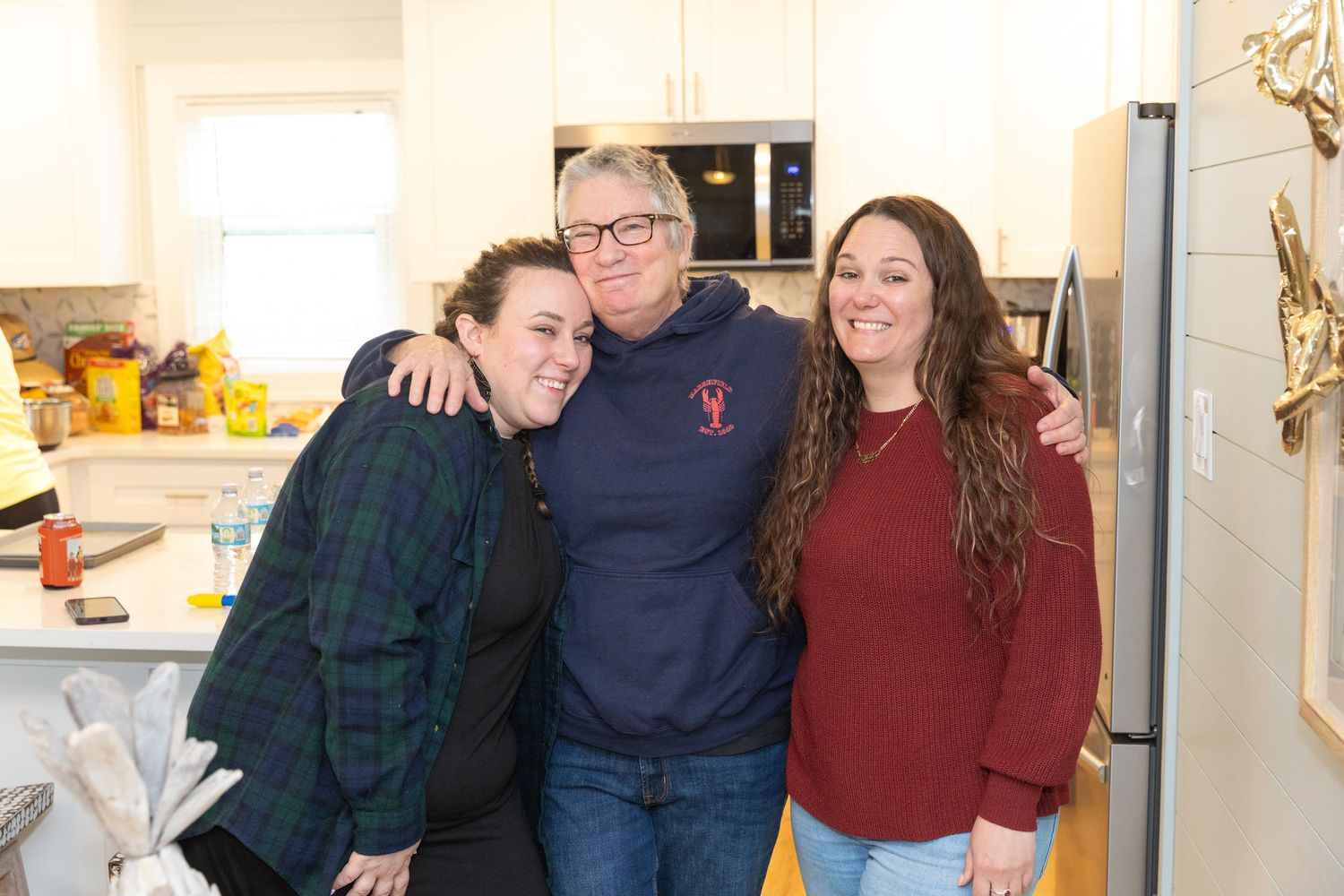 Three women smiling and hugging together in a bright modern kitchen.