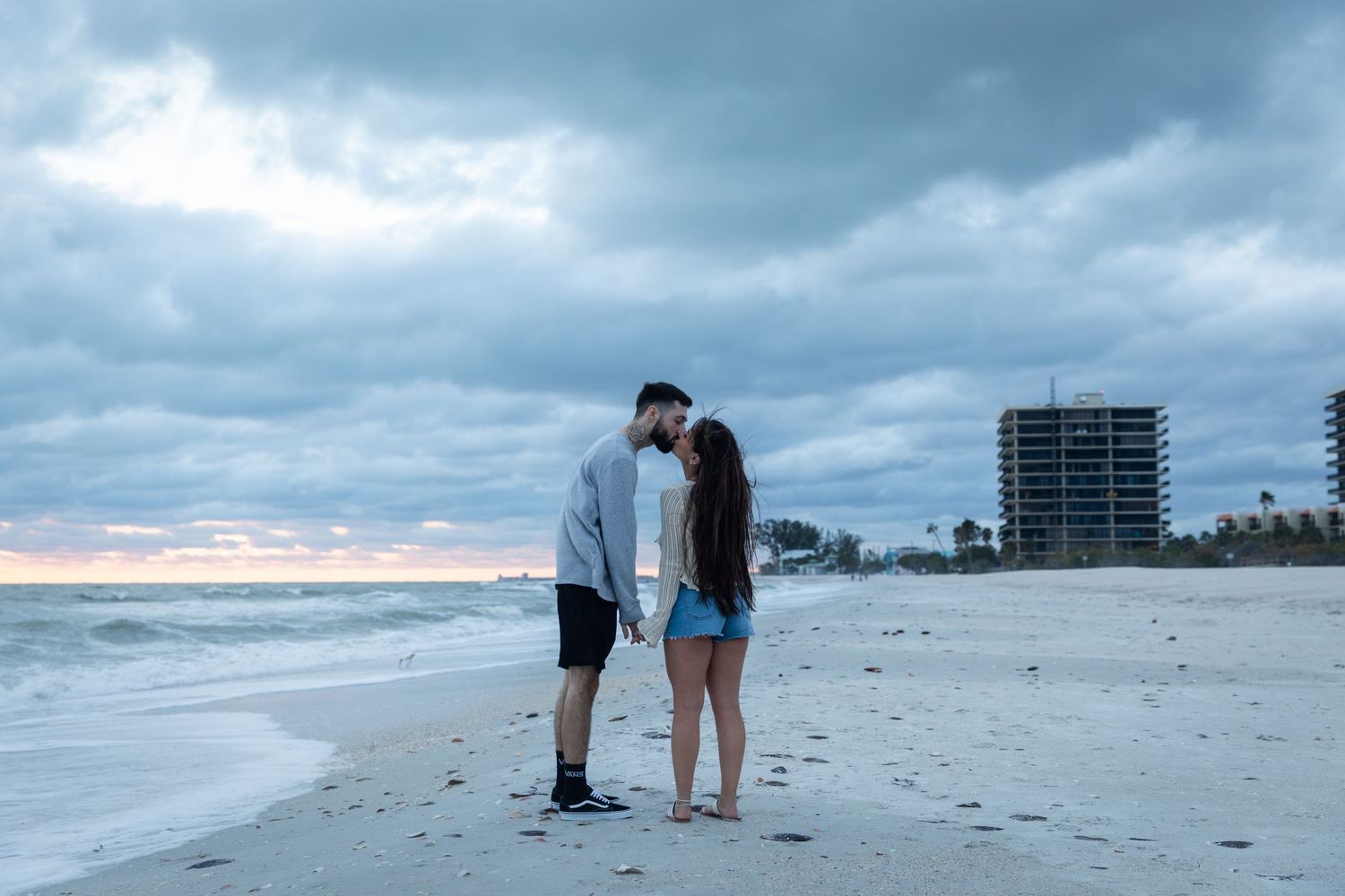 Couple sharing a kiss on a sandy beach at dusk with ocean waves and city skyline.