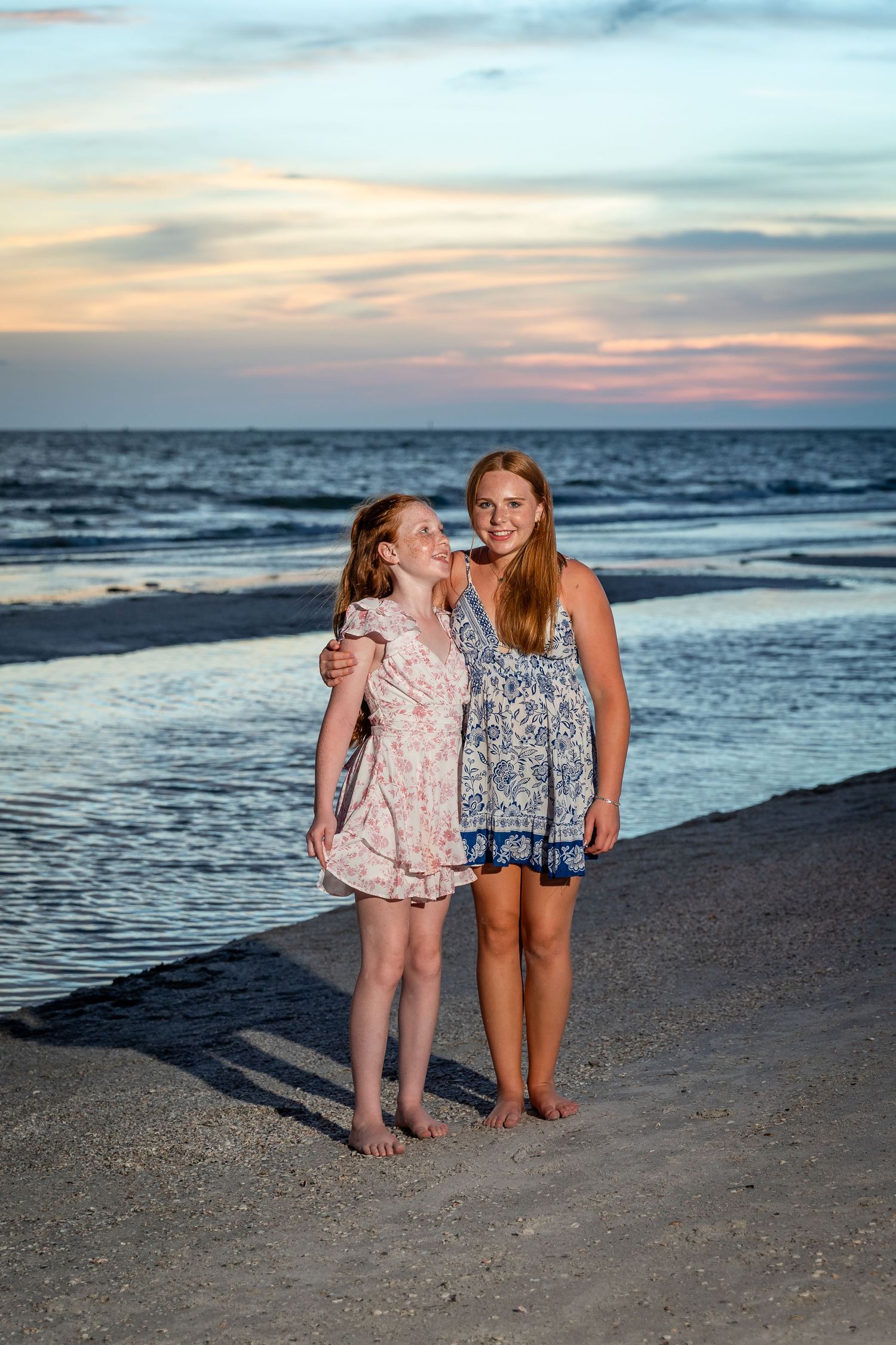 Two girls in summer dresses embrace on a sandy beach at sunset with ocean waves in the background.