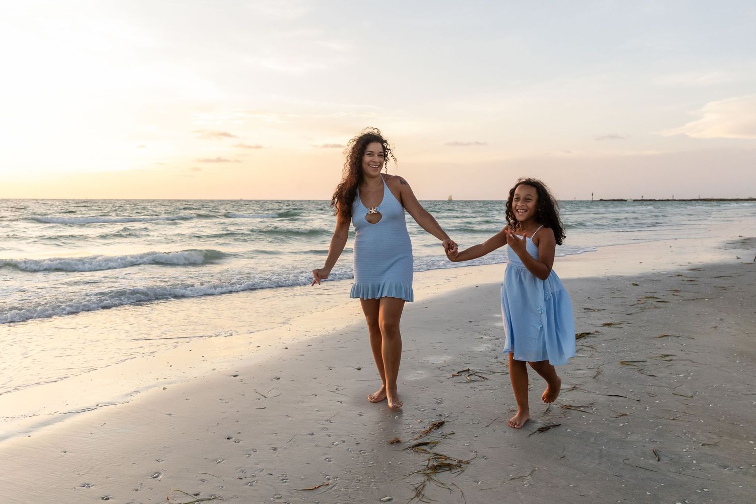 A mother and daughter in blue dresses hold hands while walking barefoot on a sandy beach at sunset.