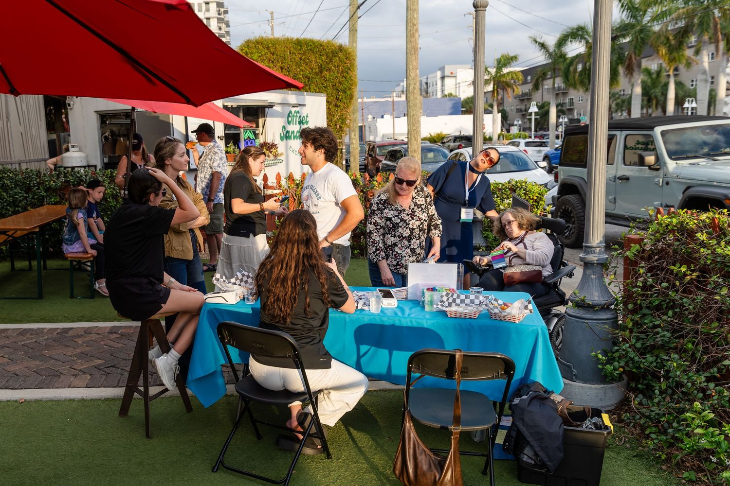 People gather around a blue table at an outdoor event in a sunny urban setting with red umbrellas and parked cars.