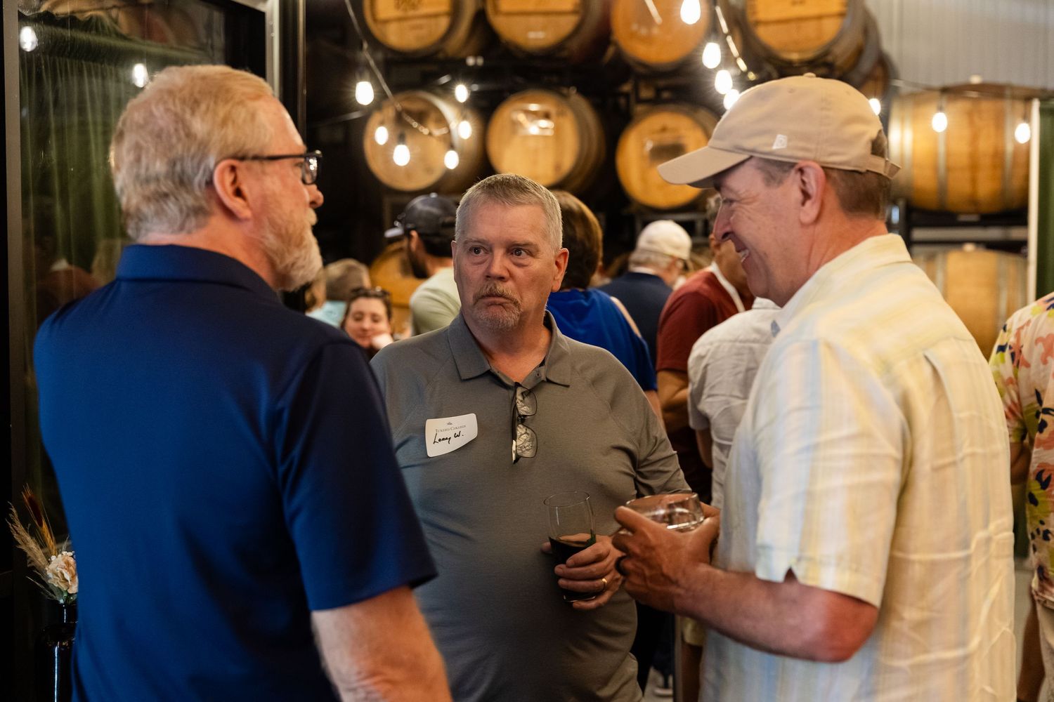 Three men engaged in conversation at a brewery networking event with barrels behind them.