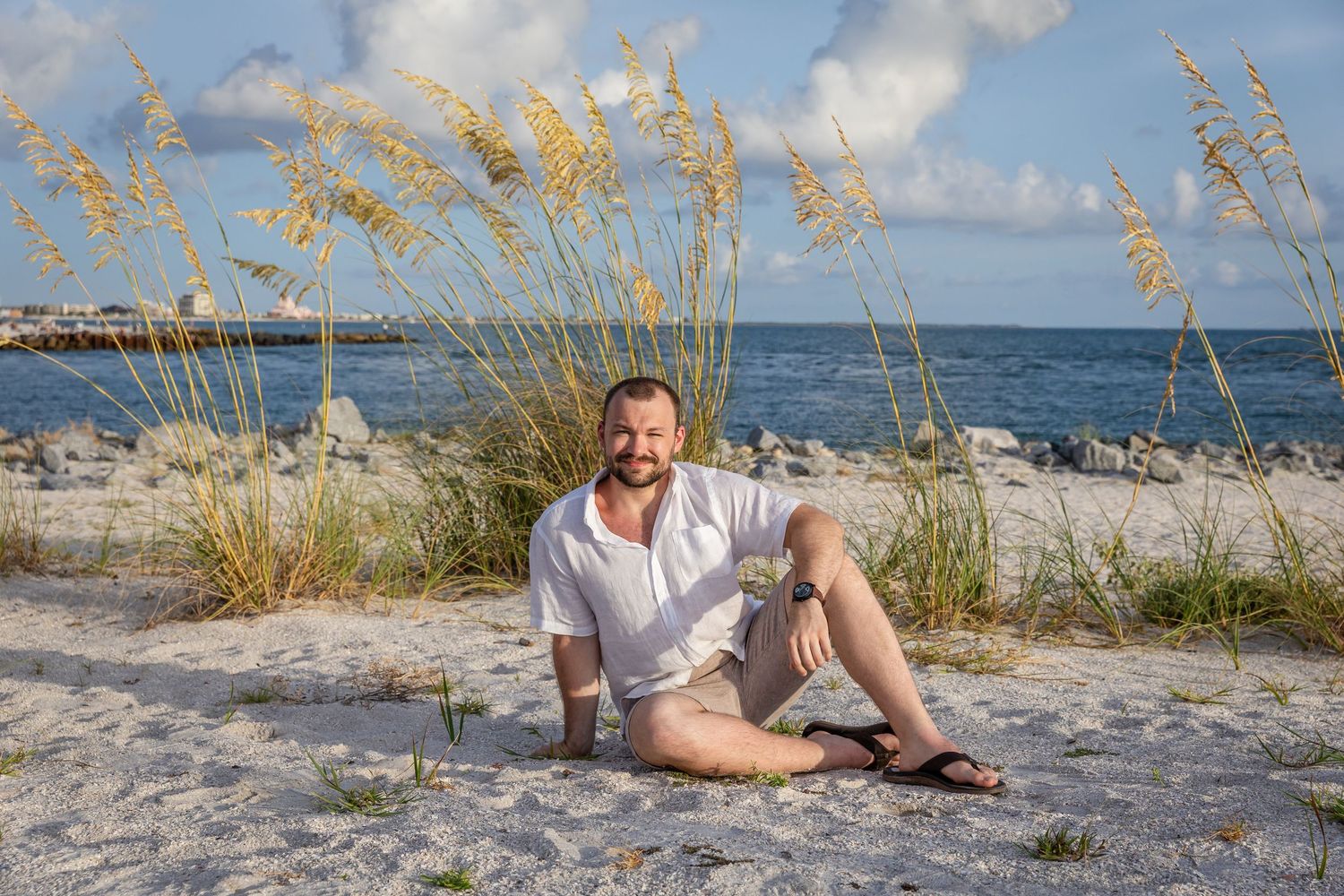 Man sitting relaxed on sandy beach with sea oats and ocean pier visible in the background on a sunny day.