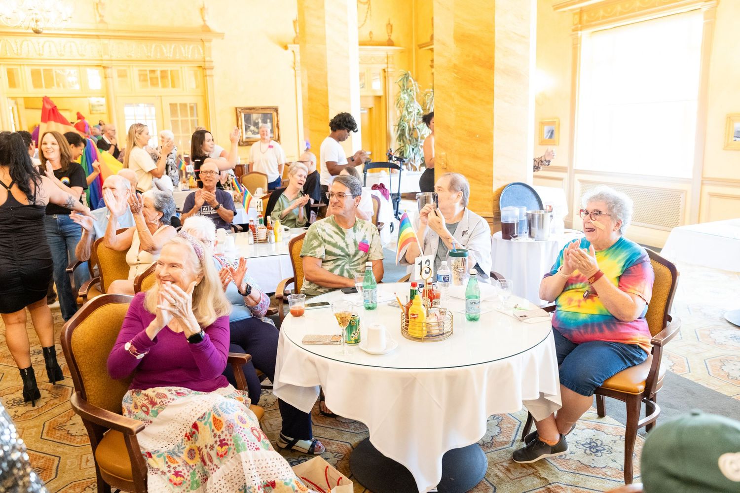 Large group of seniors seated at round tables applauding at a festive indoor celebration event.