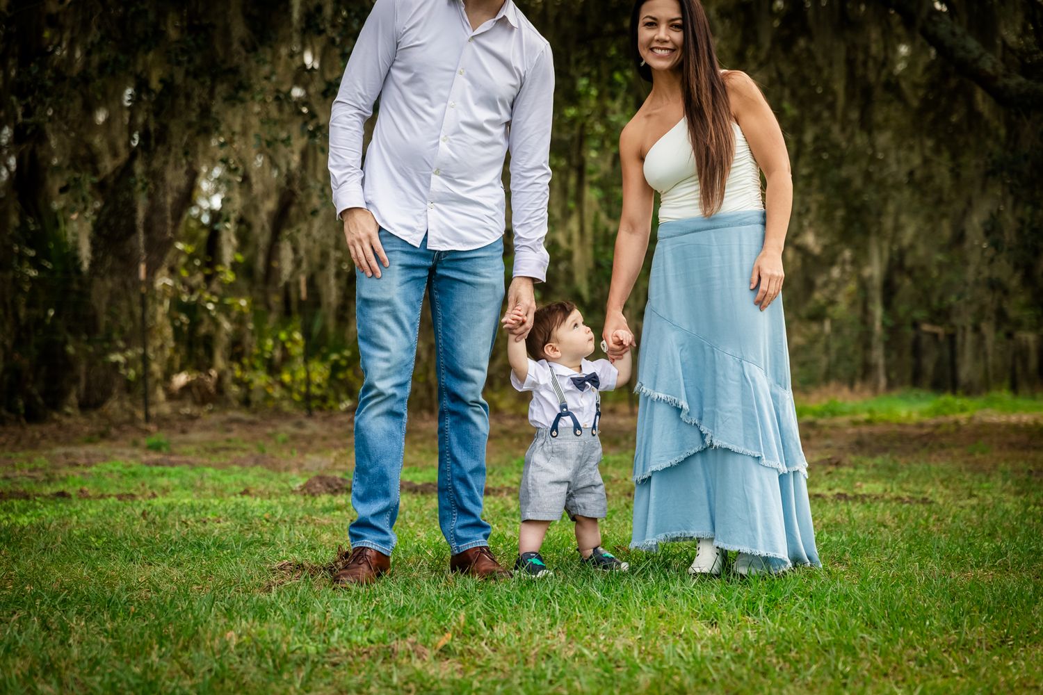 Family of three posing outdoors, parents holding toddler boy's hands on green grass with moss-covered trees behind them.