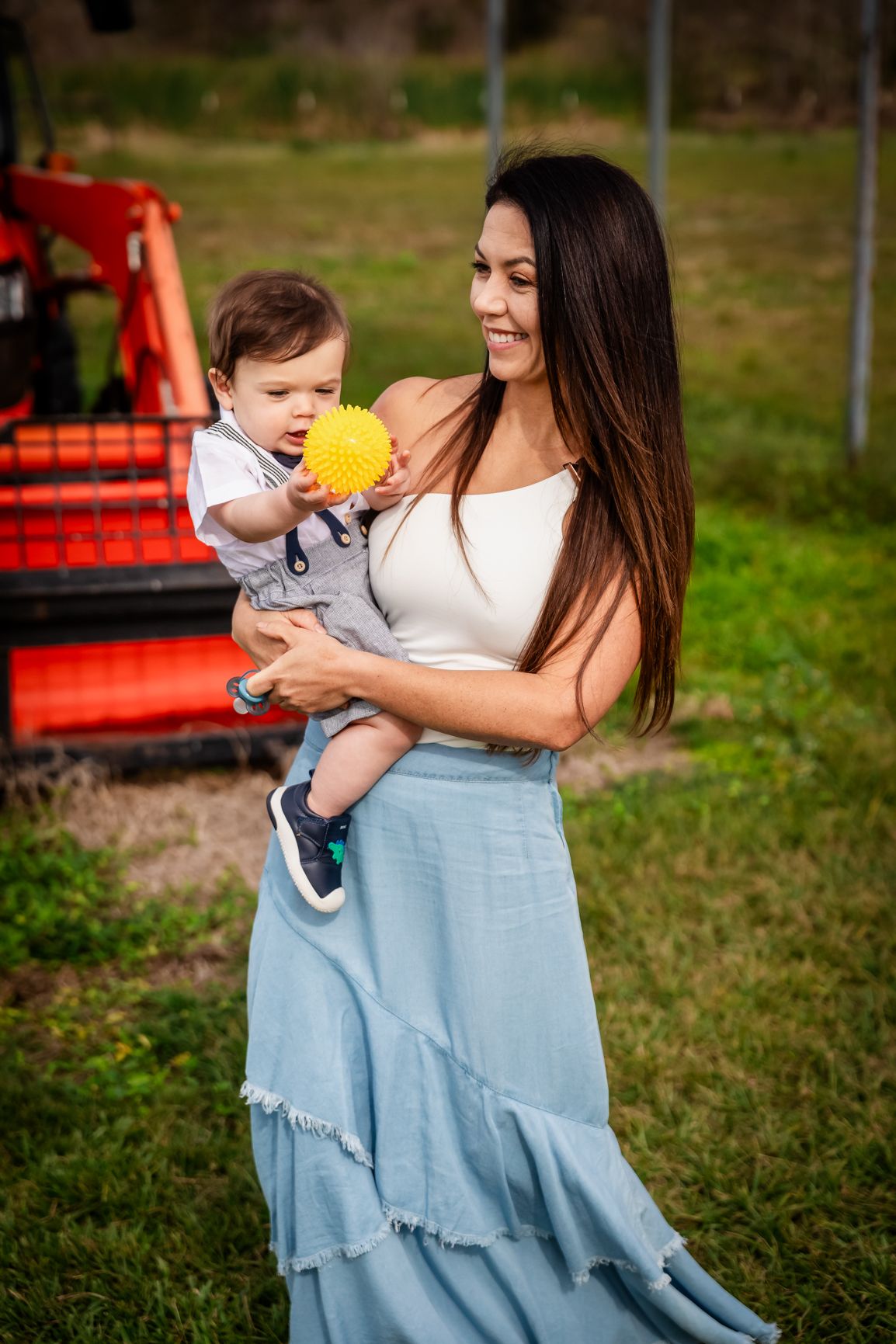 Loving mother in blue skirt holding baby playing with yellow spiky ball near red tractor outdoors.
