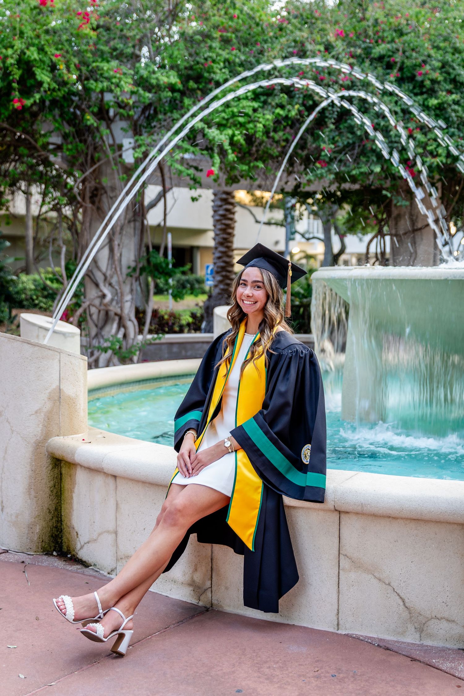 Smiling graduate in cap and gown with yellow sash poses by a fountain surrounded by lush greenery.