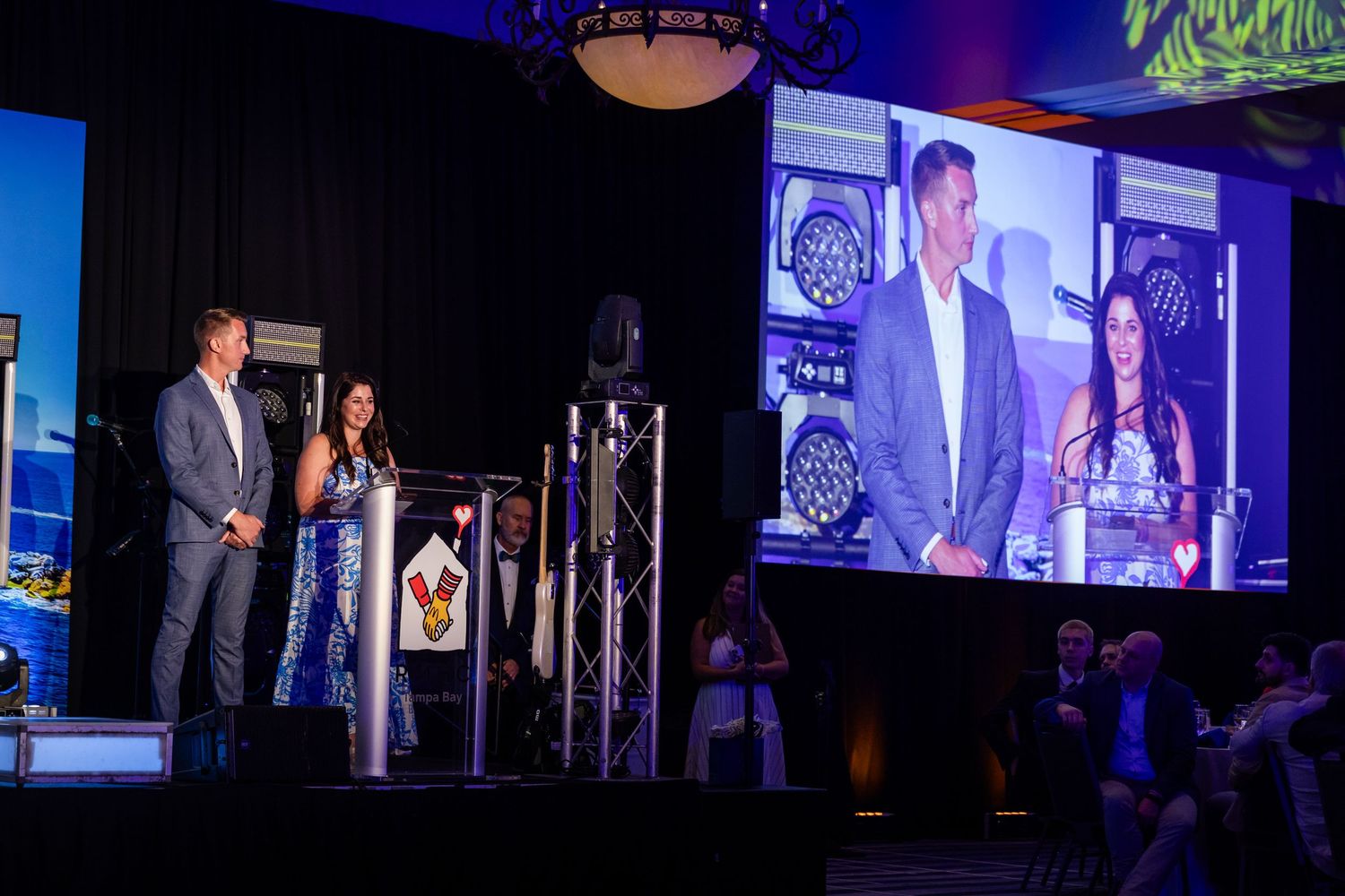 A man and woman present awards on stage at a formal gala event, with their image displayed on a large screen behind them.