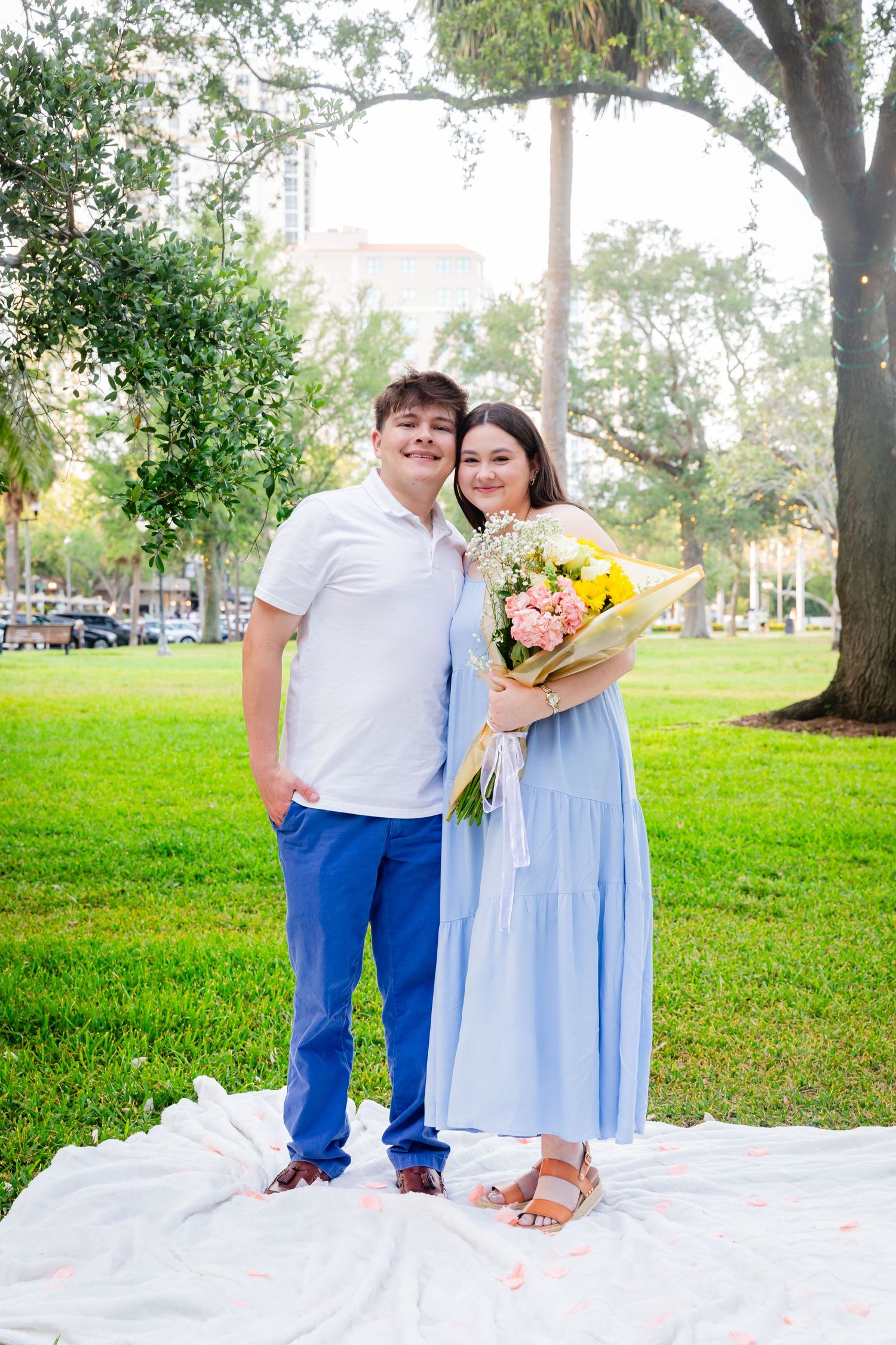 Young couple posing on white blanket in park, woman in blue dress holding colorful bouquet, man in white shirt and blue pants.