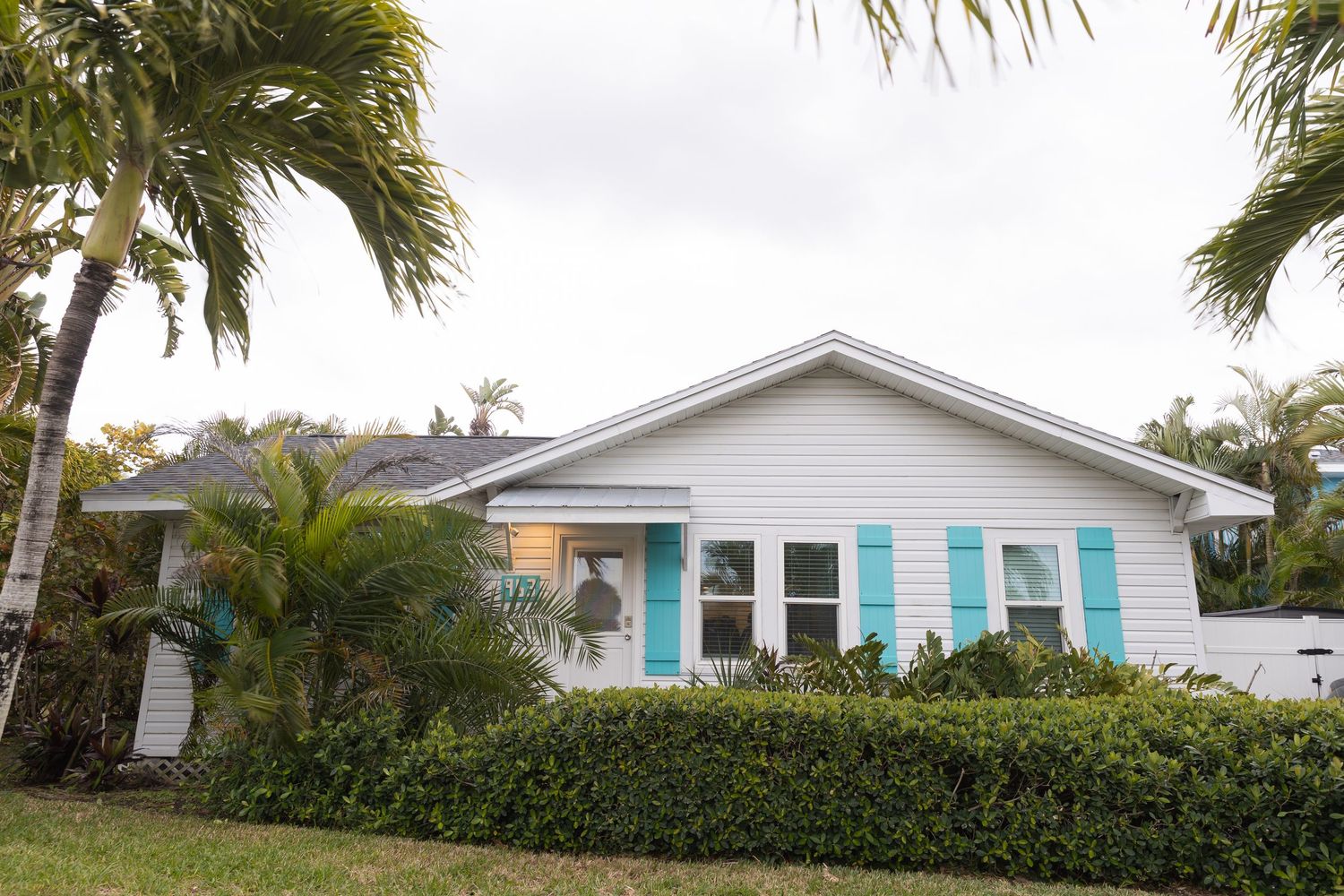 White coastal cottage home with turquoise shutters surrounded by lush palm trees and tropical landscaping.