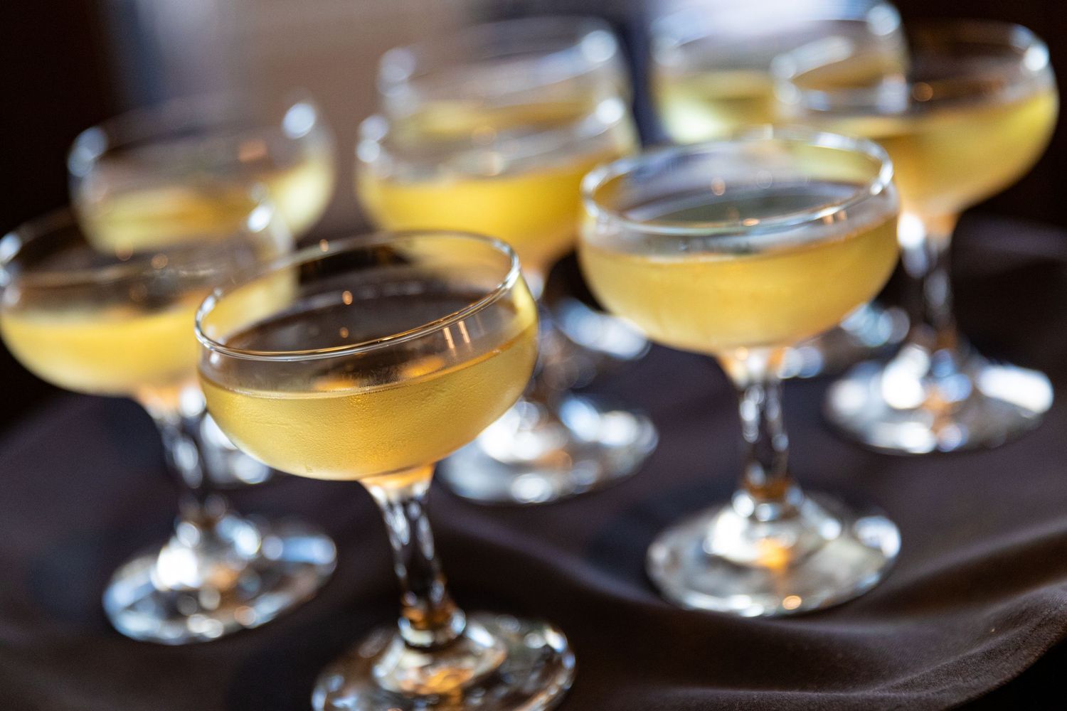 Six coupe glasses filled with yellow cocktails arranged on a dark tray at an elegant event.