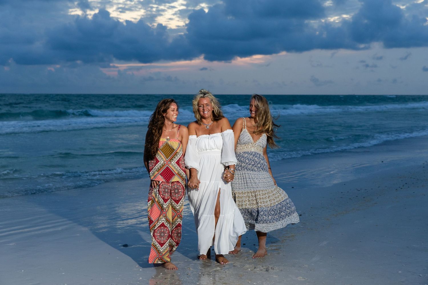 Three women in elegant dresses walk along a beach shoreline with turquoise waves at dusk.