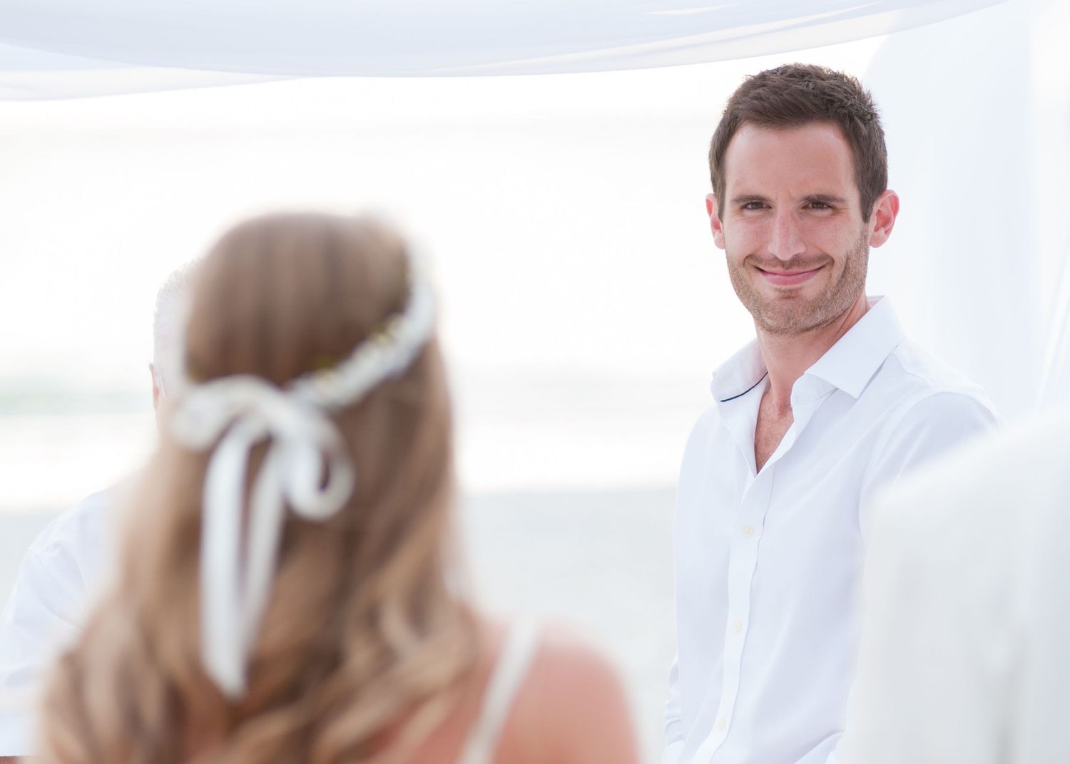 A groom in a white shirt smiles at his bride during an outdoor beach wedding ceremony.