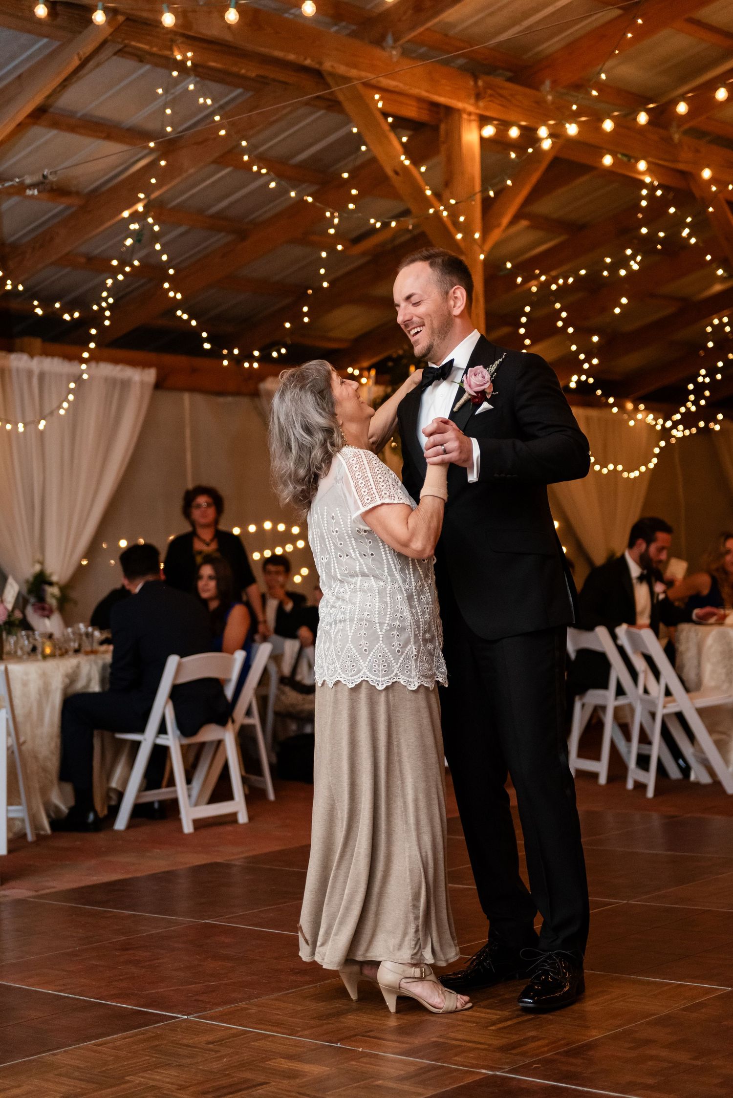 Groom in black tuxedo shares mother-son dance under warm string lights at rustic barn reception.