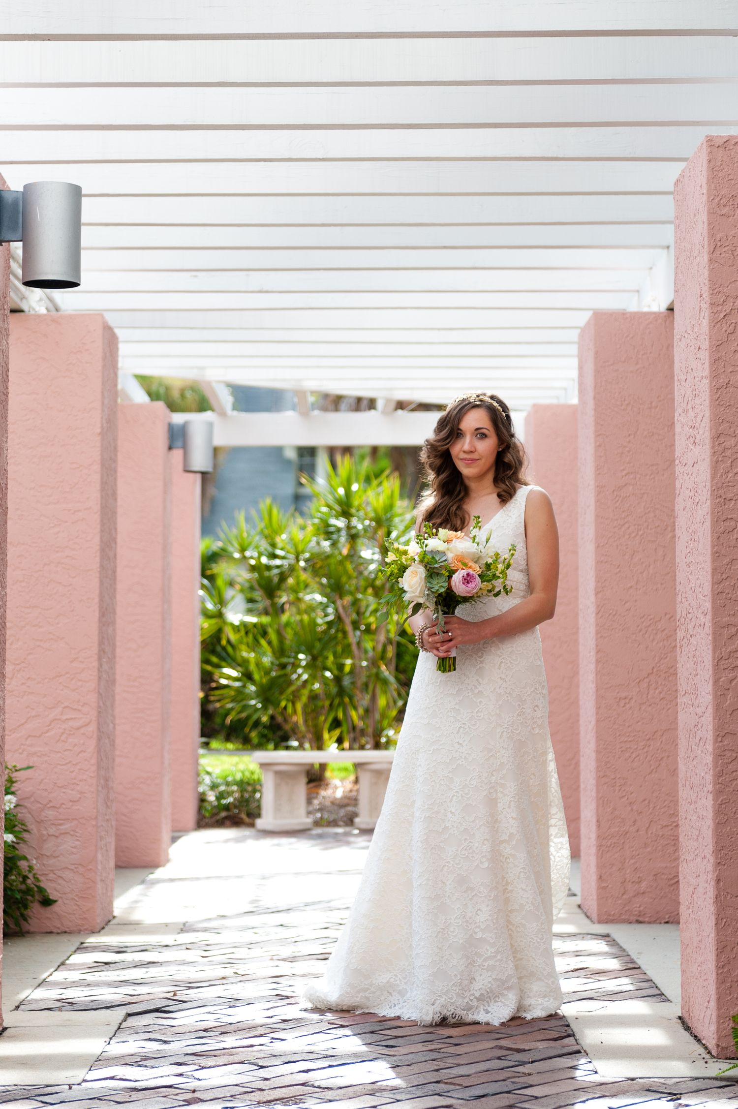 Bride in white lace gown holding bouquet standing between pink columns in outdoor walkway at The Vinoy Resort.