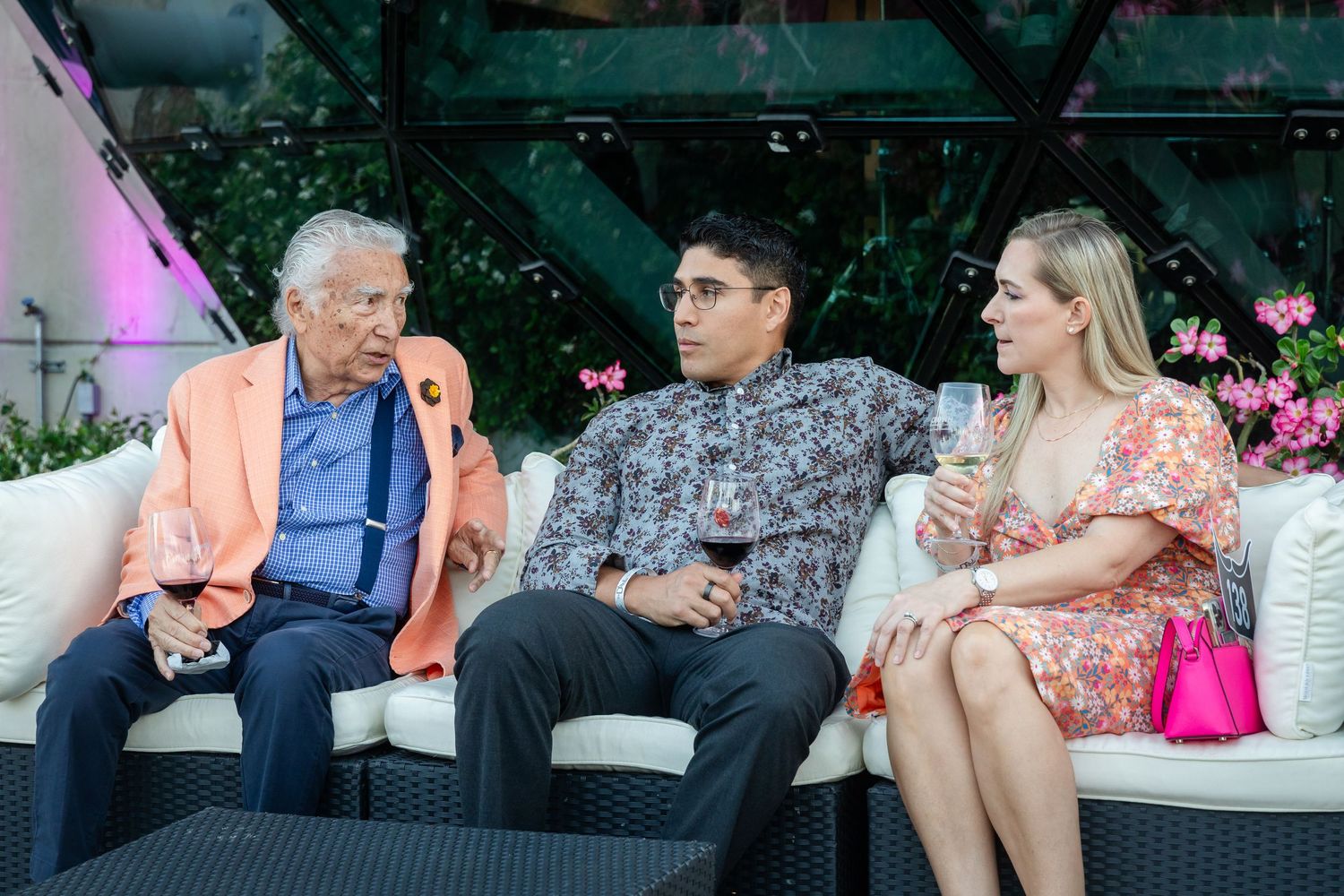 Three guests enjoying drinks and conversation on a white sofa at an elegant indoor garden event.