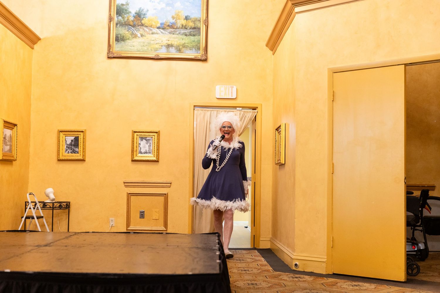 Performer in a navy and white costume stands in an ornate yellow-walled venue near a small stage.