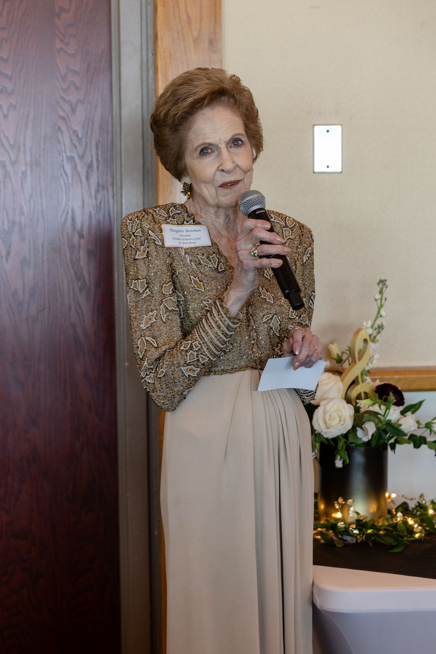 Elegant senior woman in gold sequined jacket speaks into microphone at formal Florida Orchestra fundraising event.