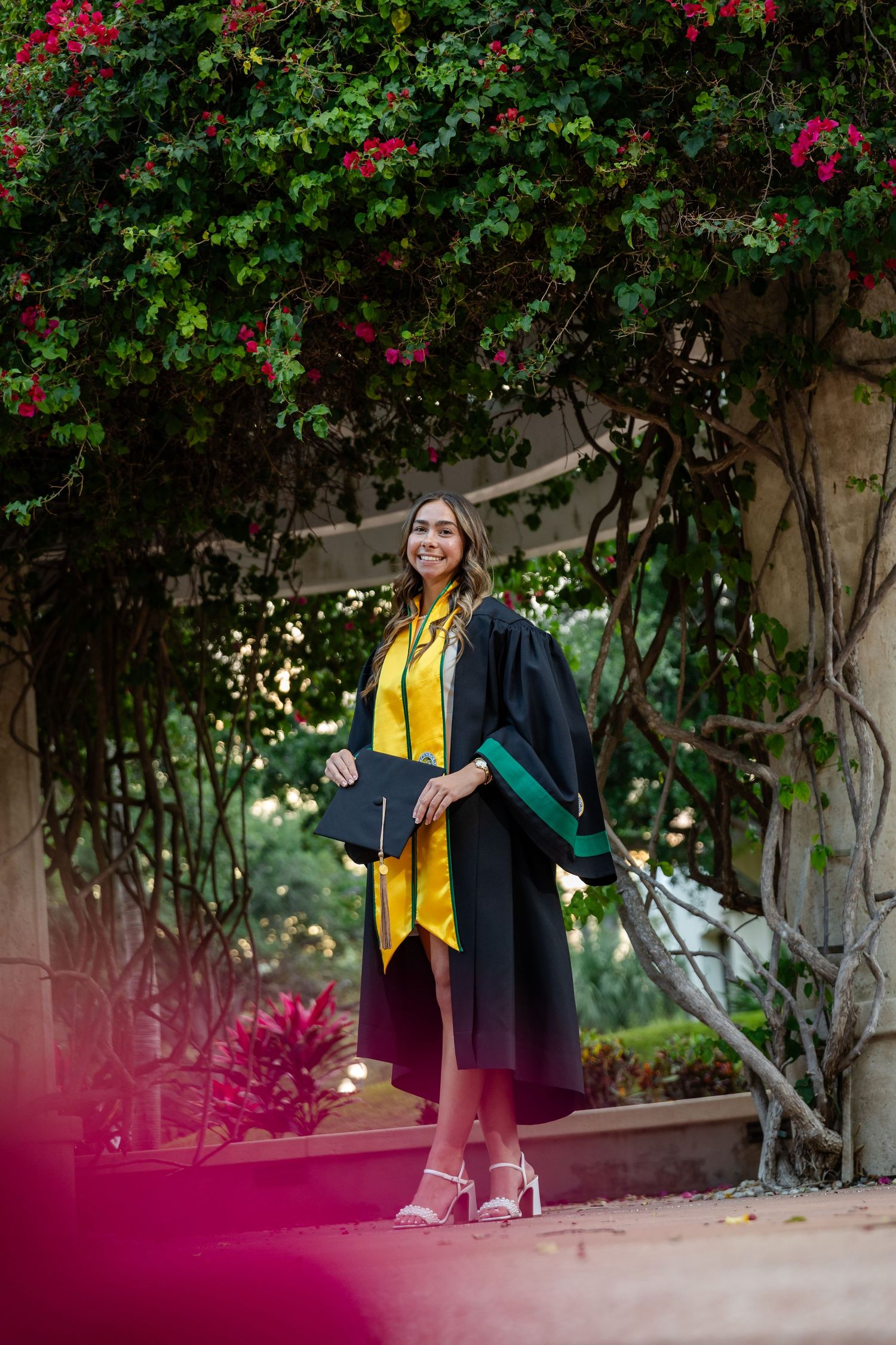Young woman in black graduation gown with yellow stole smiles outdoors under flowering vines.