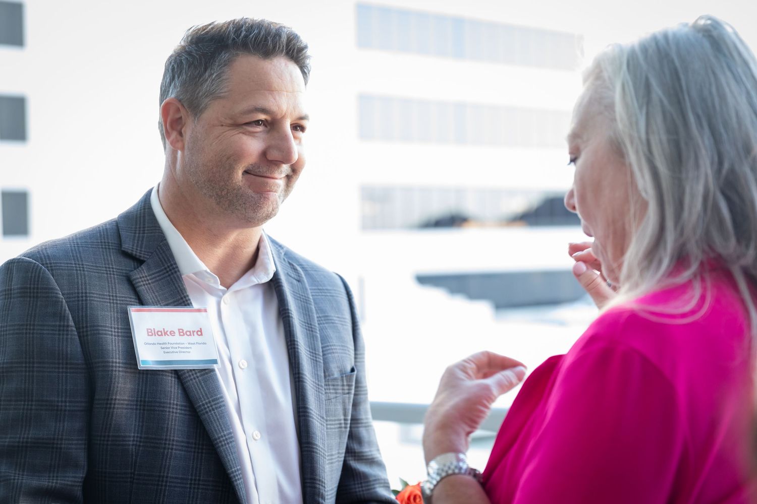 A smiling man wearing a name badge converses with a woman in pink at a professional networking event.