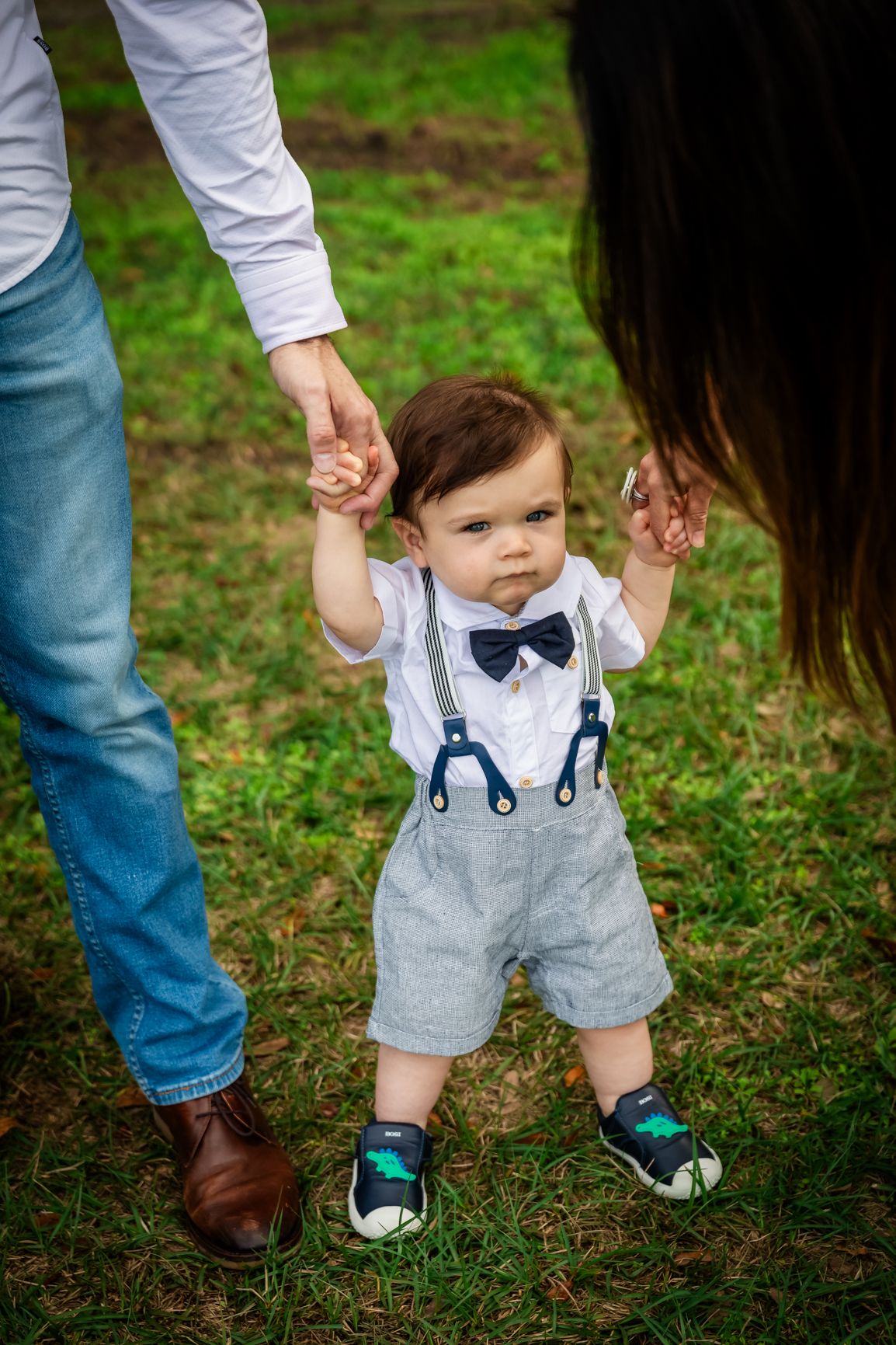 Toddler boy in suspenders and bow tie taking first steps outdoors, held by parents on green grass.