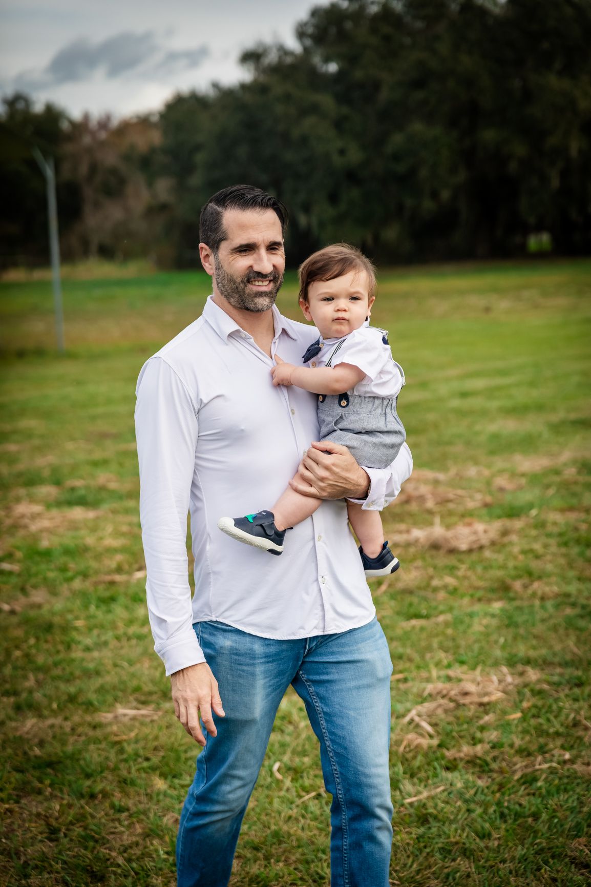 A smiling man in a white shirt and jeans holds a baby boy in a gray outfit in a green park.