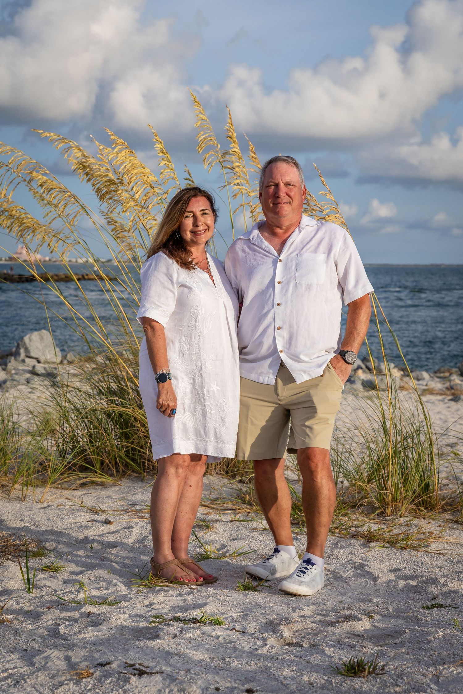 Smiling couple in white linen outfits posing on sandy beach with sea oats and ocean backdrop.