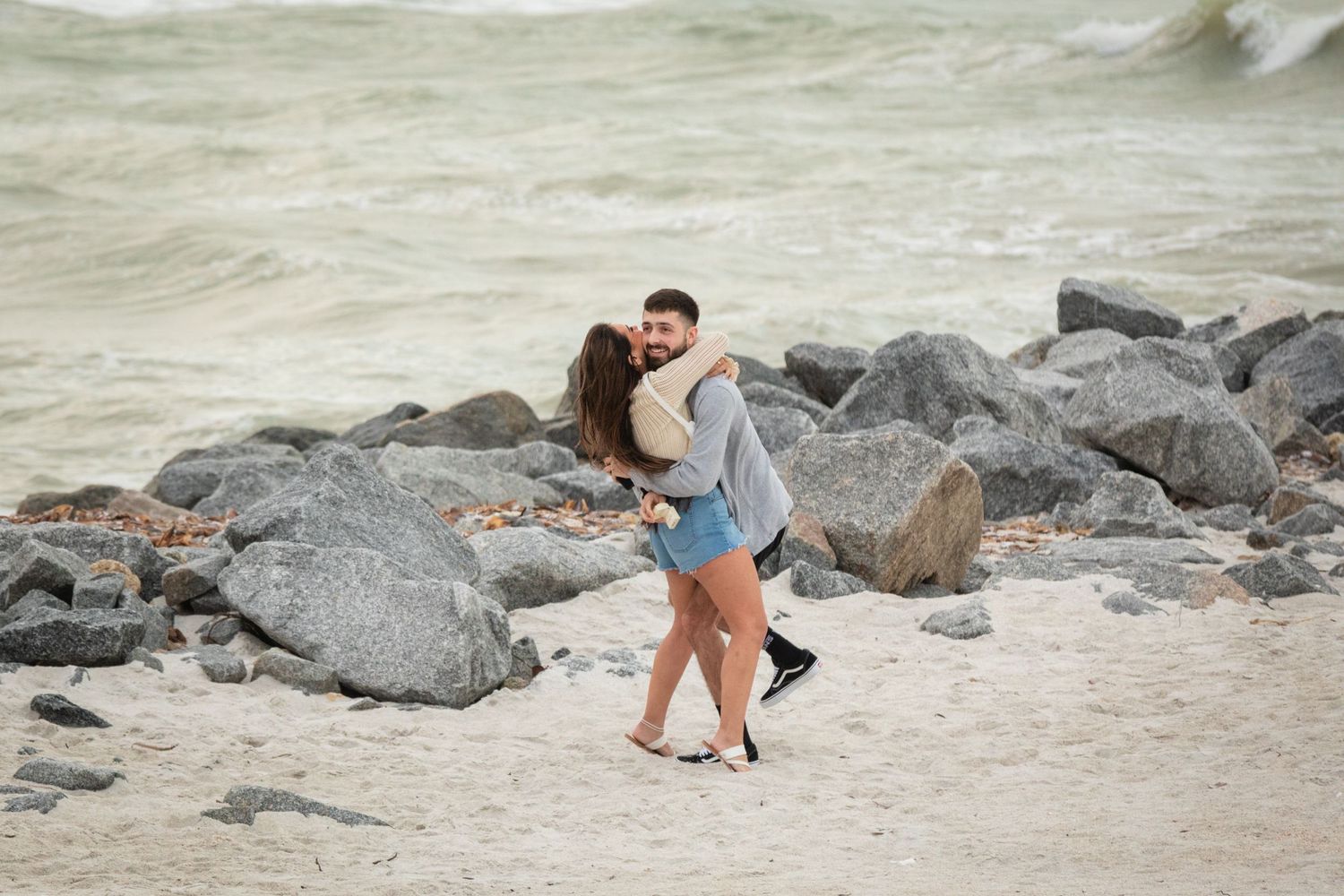 A couple embracing and dancing romantically on a sandy beach with rocky shoreline and ocean waves behind them.