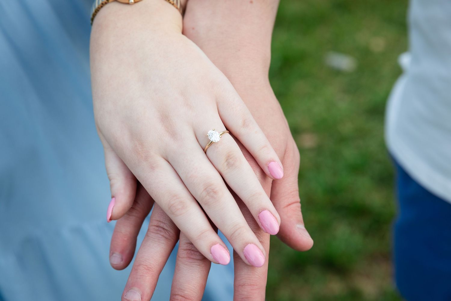 Two hands stacked together displaying an engagement ring with a pearl stone and pink manicured nails outdoors.