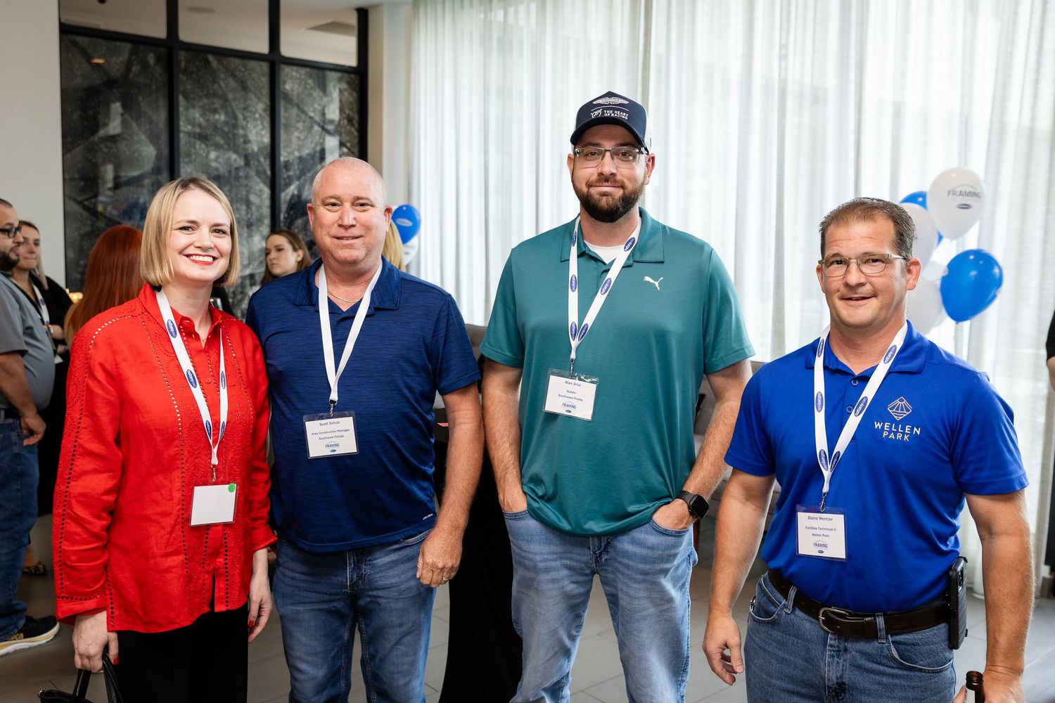 Four professionals wearing lanyards smile together at a corporate event with blue balloons in the background.