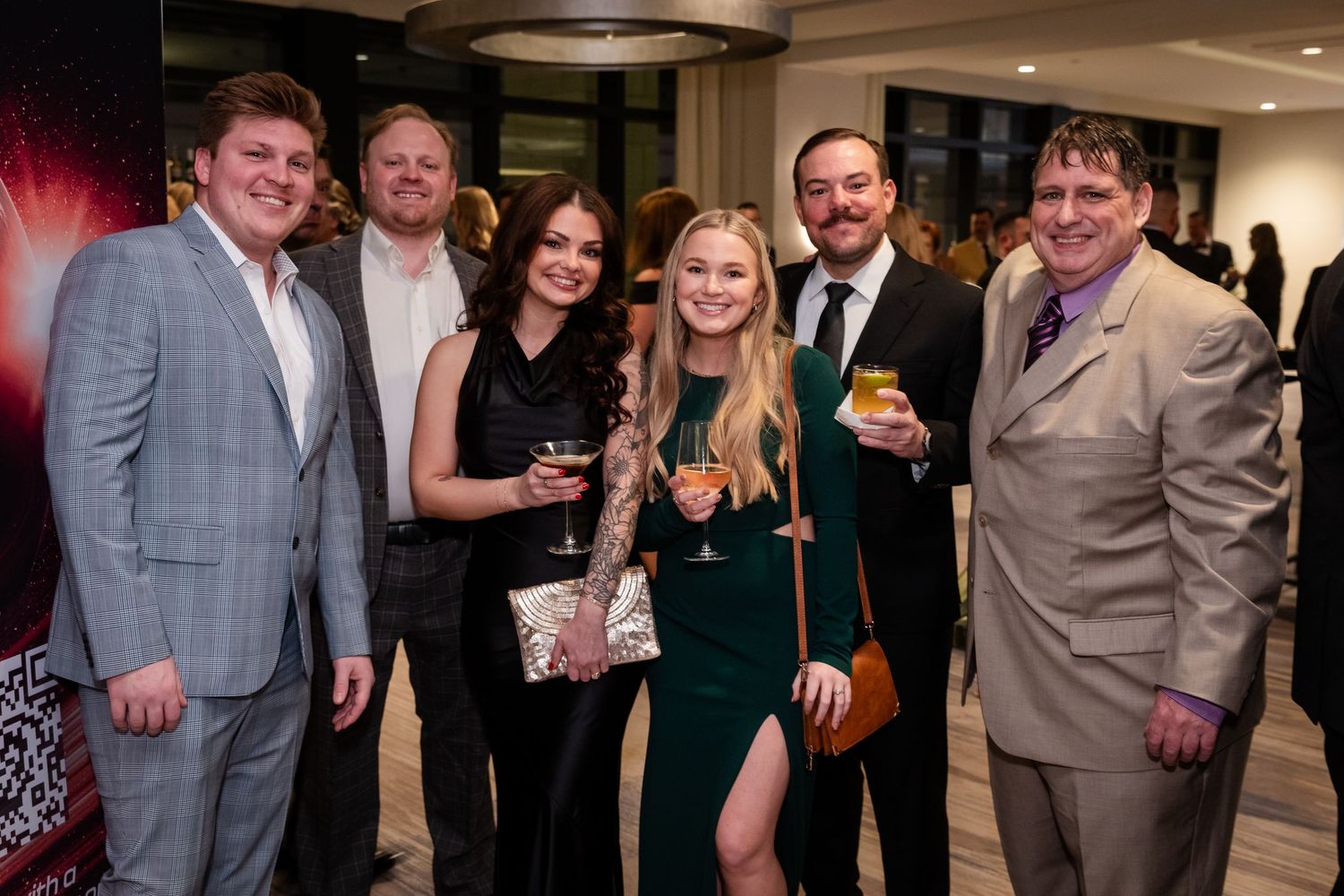 Group of seven adults posing together at a formal evening event, smiling and holding drinks in elegant attire.