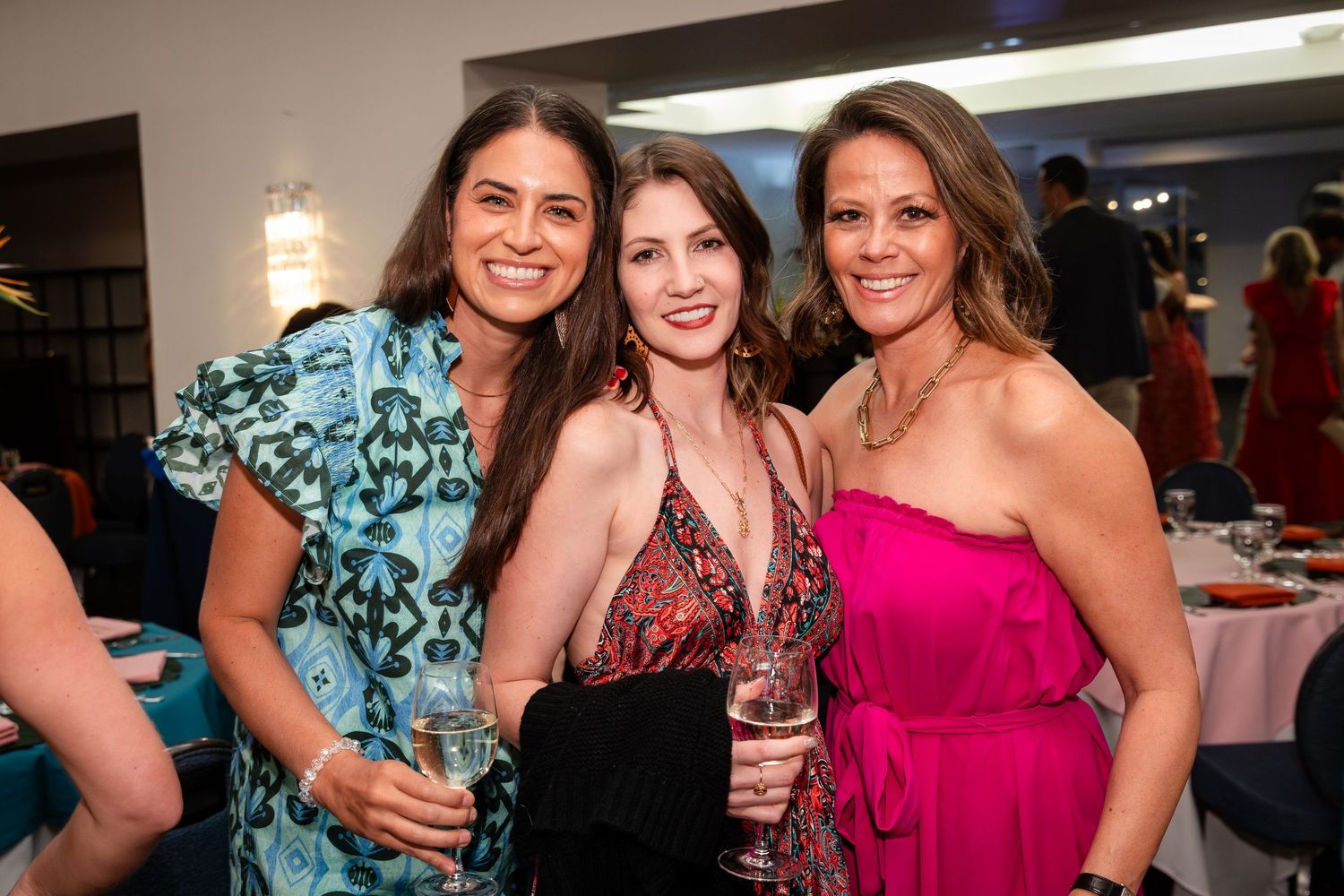 Three women smiling and holding drinks at an elegant indoor social event, wearing colorful dresses.