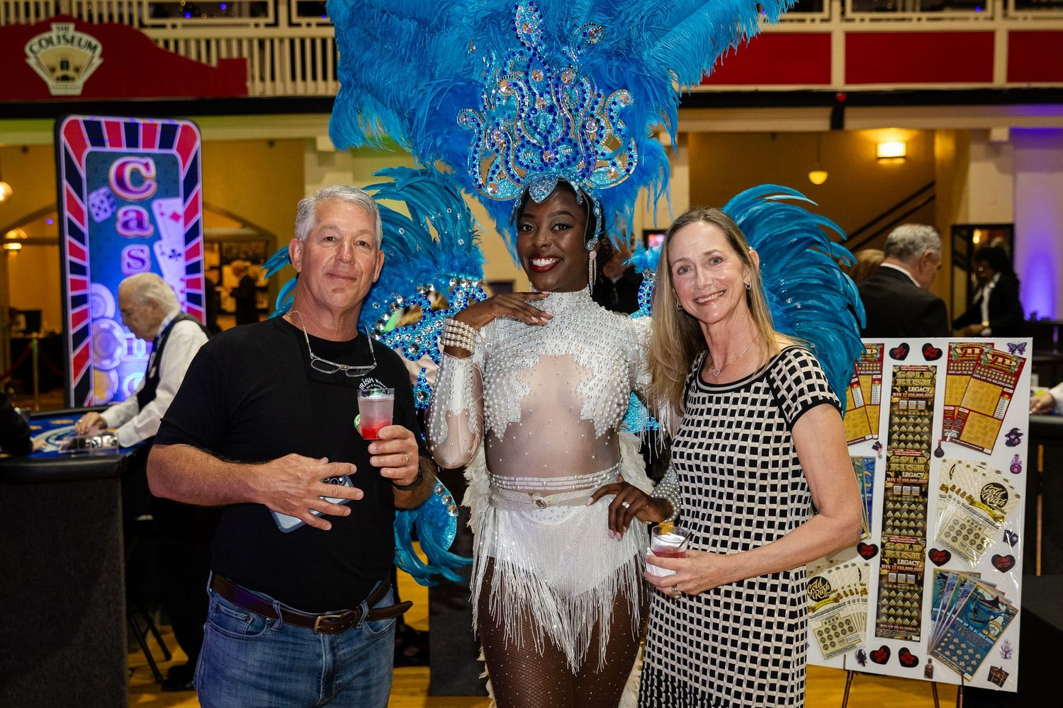 Three people pose together at a carnival-themed event, with a performer in colorful blue feathered costume.