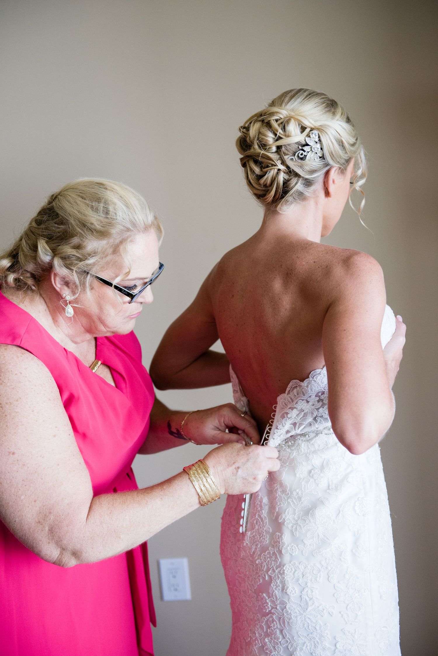Mother in pink dress helping bride button up lace wedding gown during bridal preparations indoors.