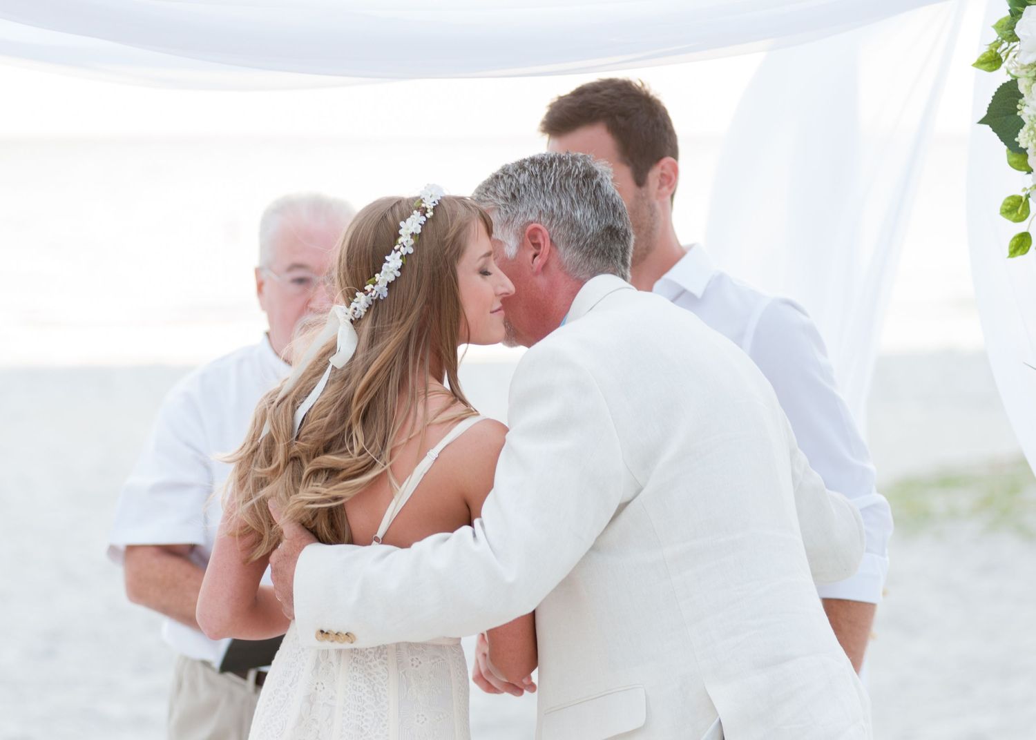 Couple sharing a first kiss at a romantic beach wedding ceremony with guests and officiant in white attire.