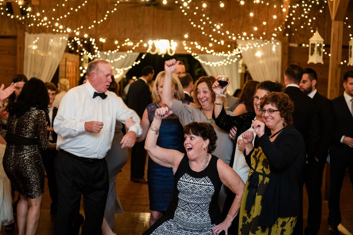 Joyful wedding guests dancing and celebrating under warm string lights at indoor barn reception.