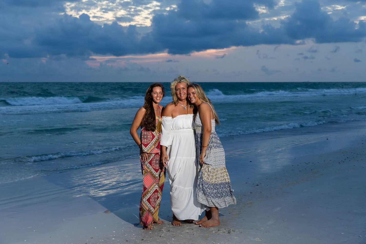 Three women pose together smiling on a sandy beach wearing maxi dresses at sunset.