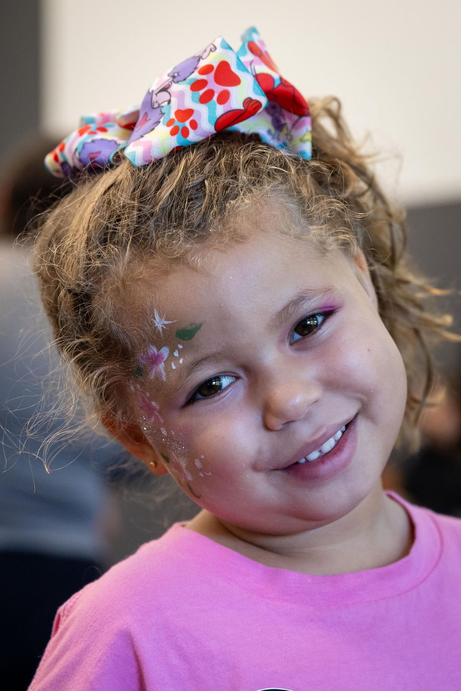 Smiling young girl with face paint and colorful bow in hair wearing a pink shirt at a fun event.
