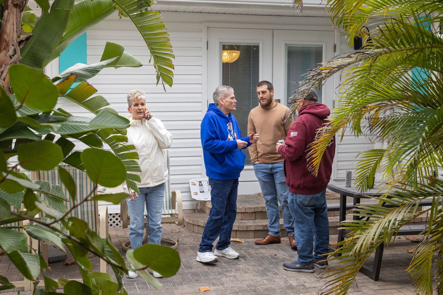 Four people having a conversation outside a white house surrounded by tropical plants and palm trees.