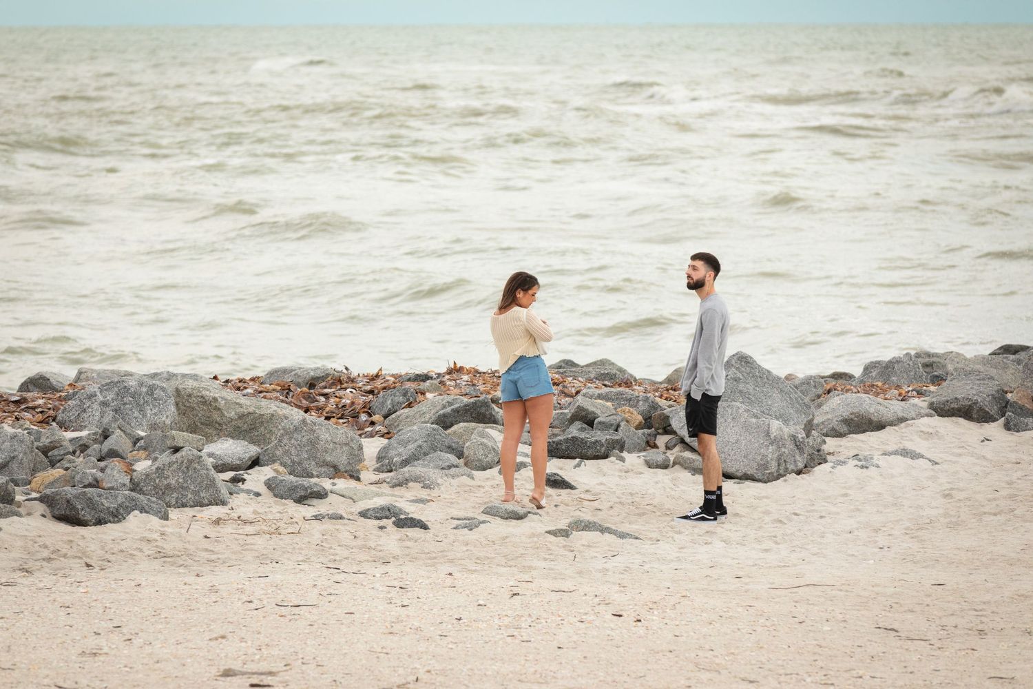Couple standing on a rocky beach near the ocean, woman in denim shorts facing a bearded man in gray shirt.