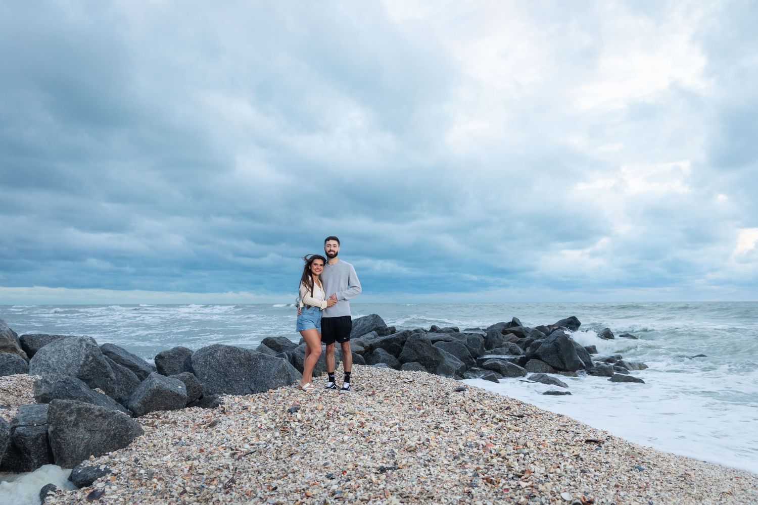 Couple standing on rocky beach shoreline with dramatic cloudy sky and ocean waves in background.